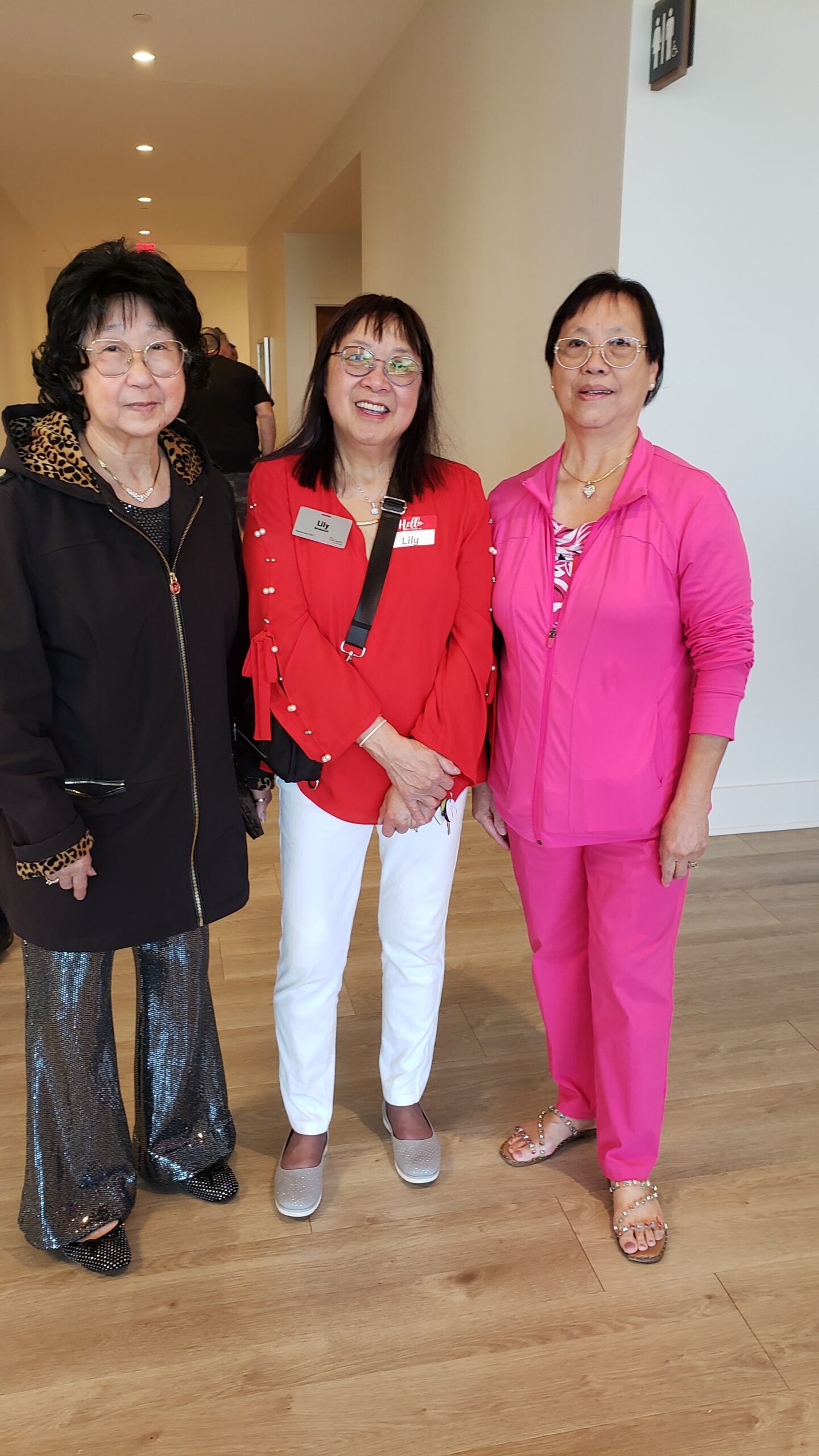 Three women pose together in a hallway, smiling for the camera: left in a black jacket with leopard trim, center in a red blouse with name tag, right in a pink outfit.