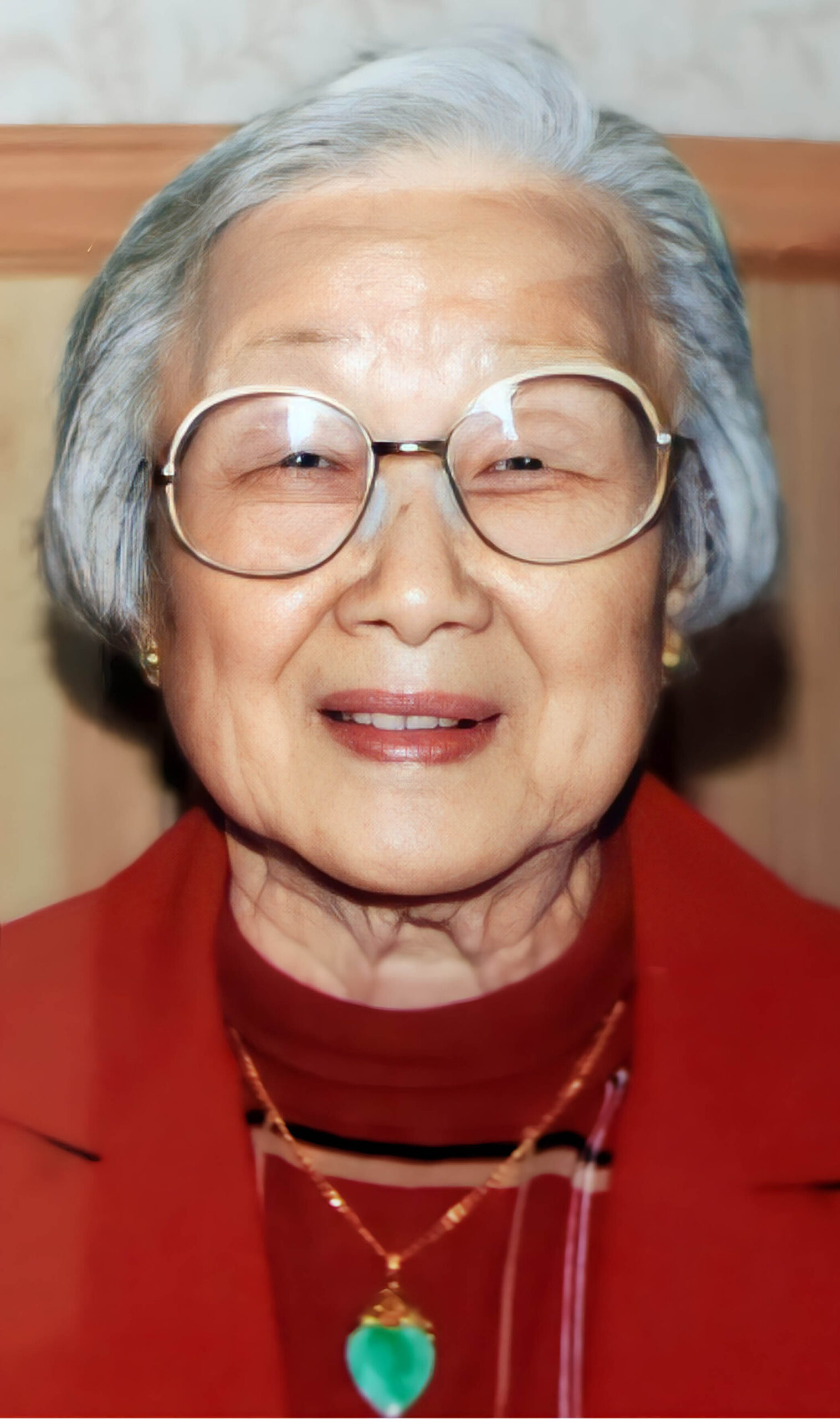 Close-up portrait of an elderly woman with short gray hair, round glasses, and a red outfit, smiling subtly with a green heart-shaped pendant on a chain visible around her neck.