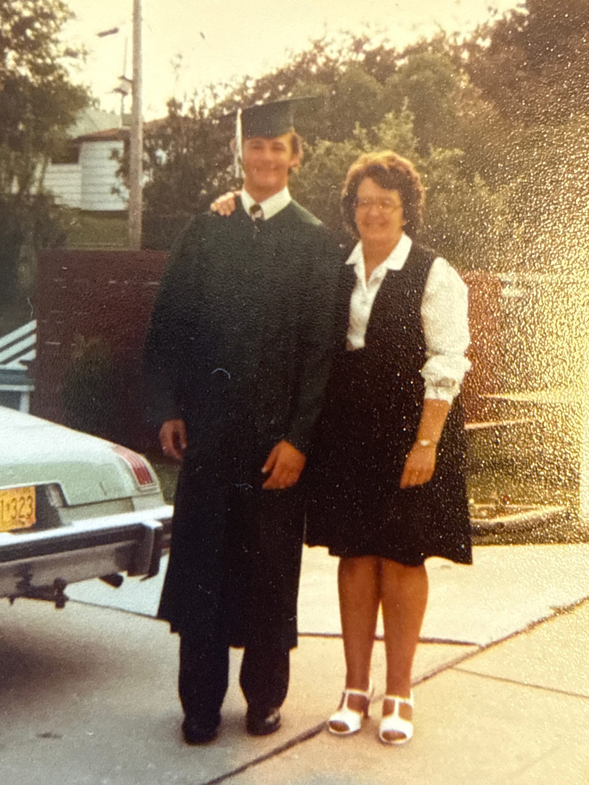 Young man in a green graduation gown and cap stands with a smiling woman in a white blouse and black vest outdoors near a parked car.