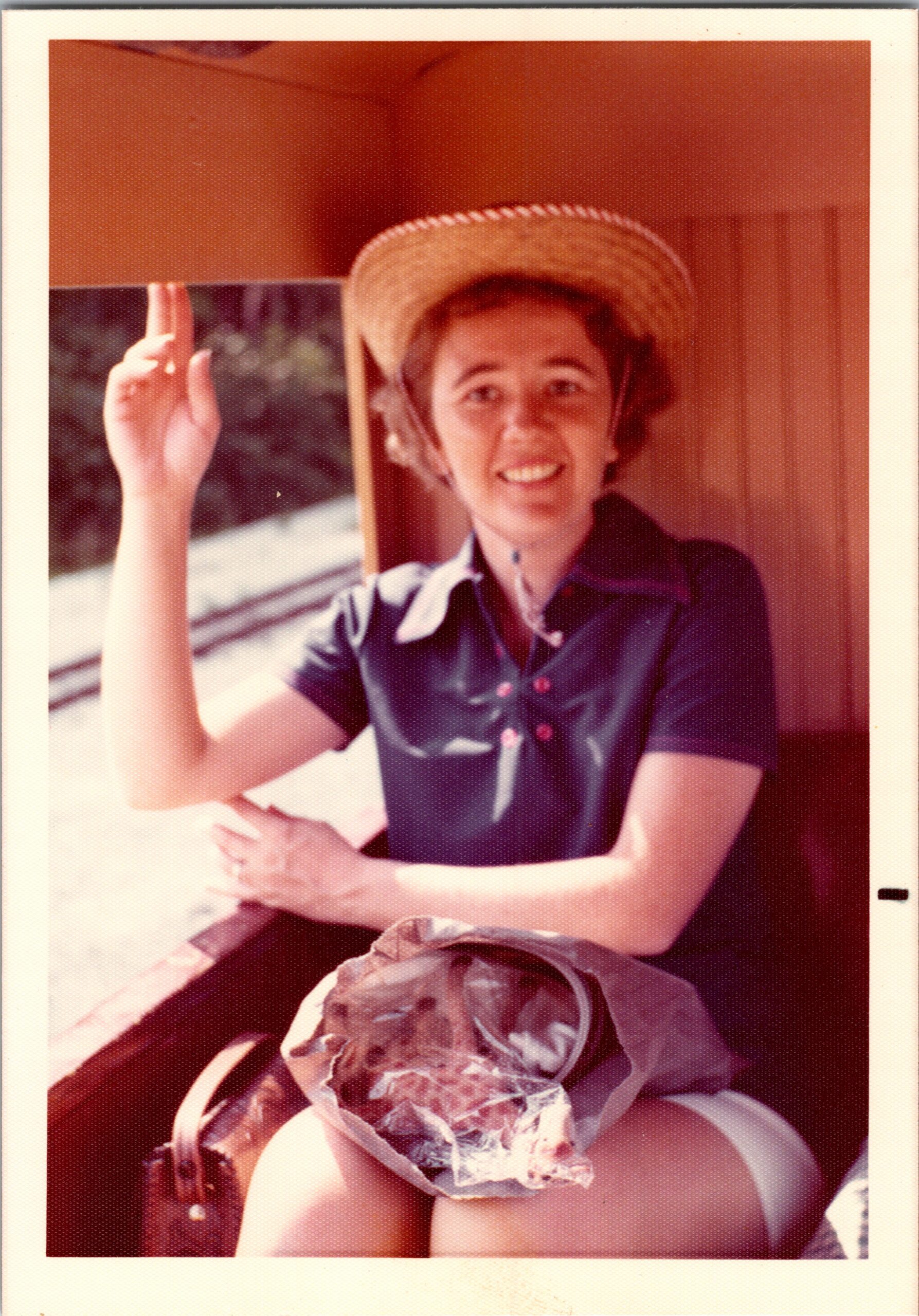 Smiling woman in a straw hat and blue shirt sits on a train, a sandwich wrapped in paper on her lap.