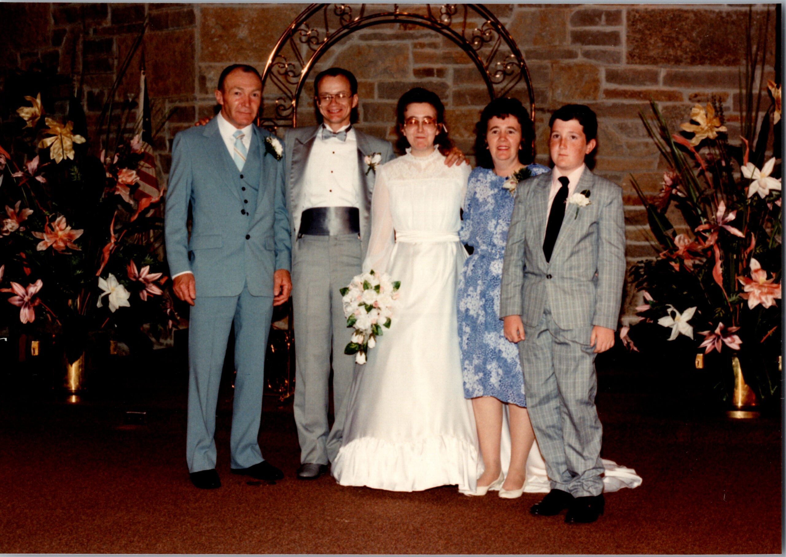 Five people pose at a wedding: the bride in a white gown flanked by two men in gray suits and two guests in blue-gray outfits, with floral arrangements in the background.