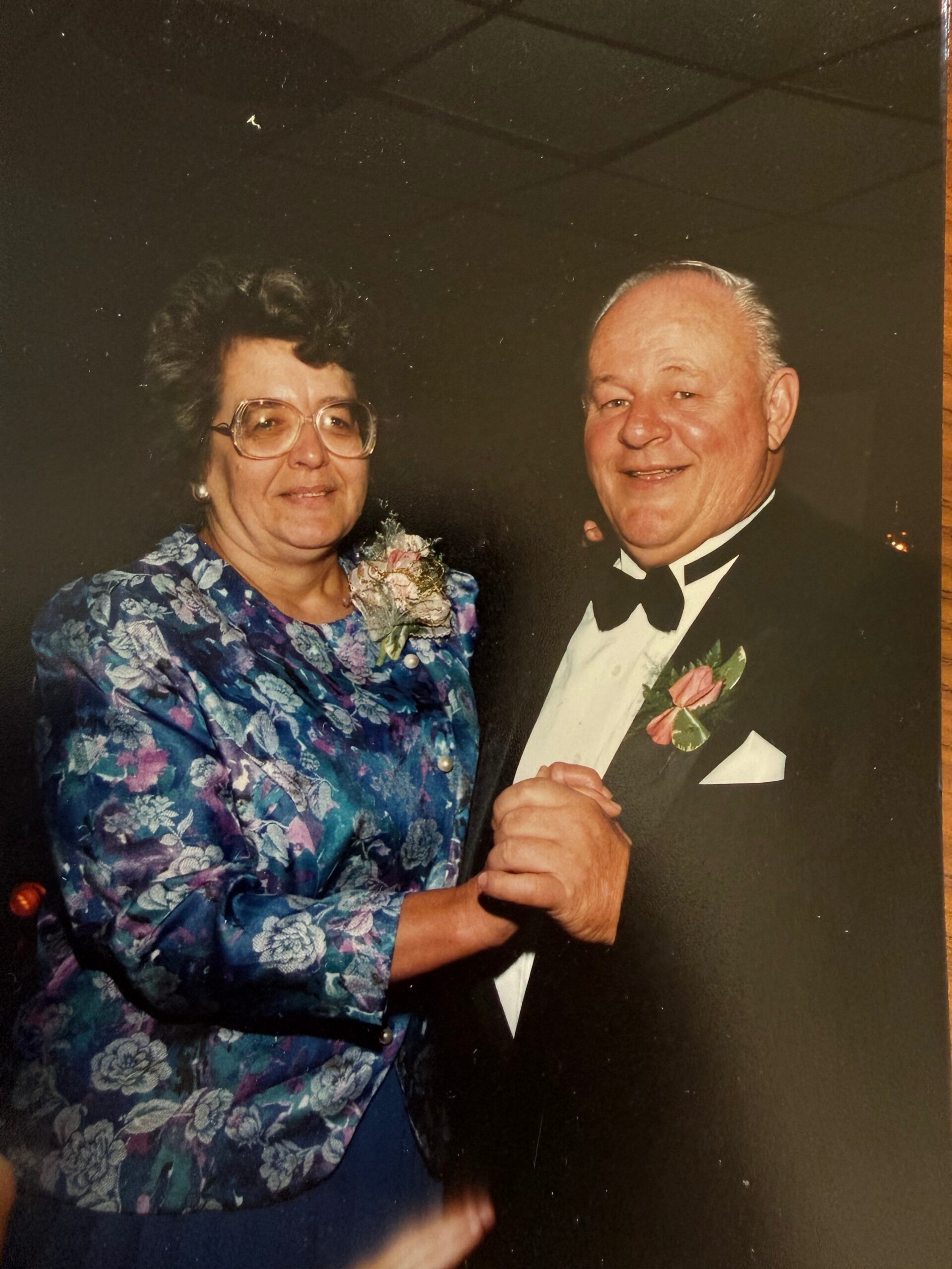 Older couple dancing together at a formal event; woman in a blue floral jacket and corsage, man in a tuxedo with boutonniere, smiling.