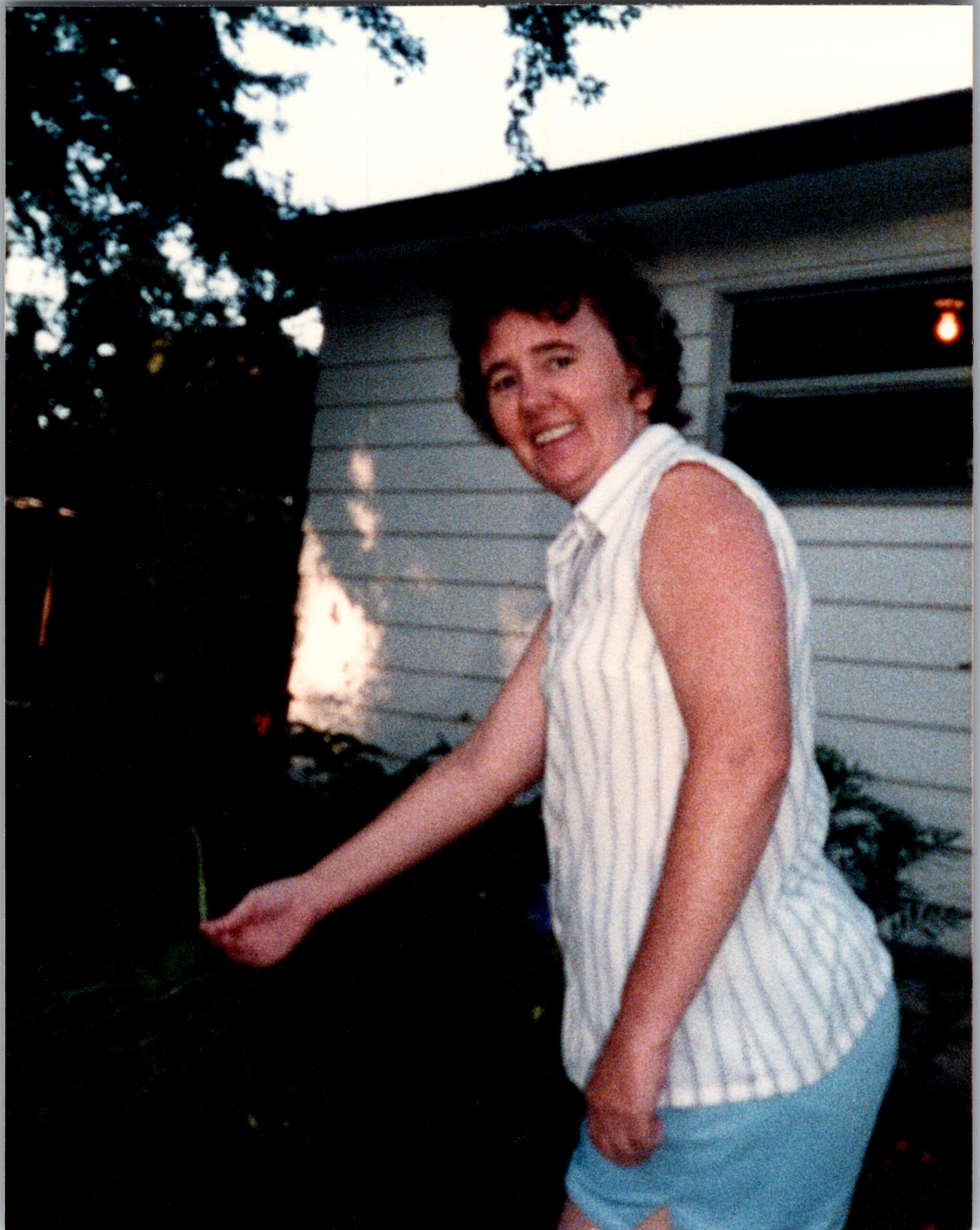 Woman with short dark hair wearing a striped sleeveless top and light blue shorts stands outside near a white house, smiling at the camera.