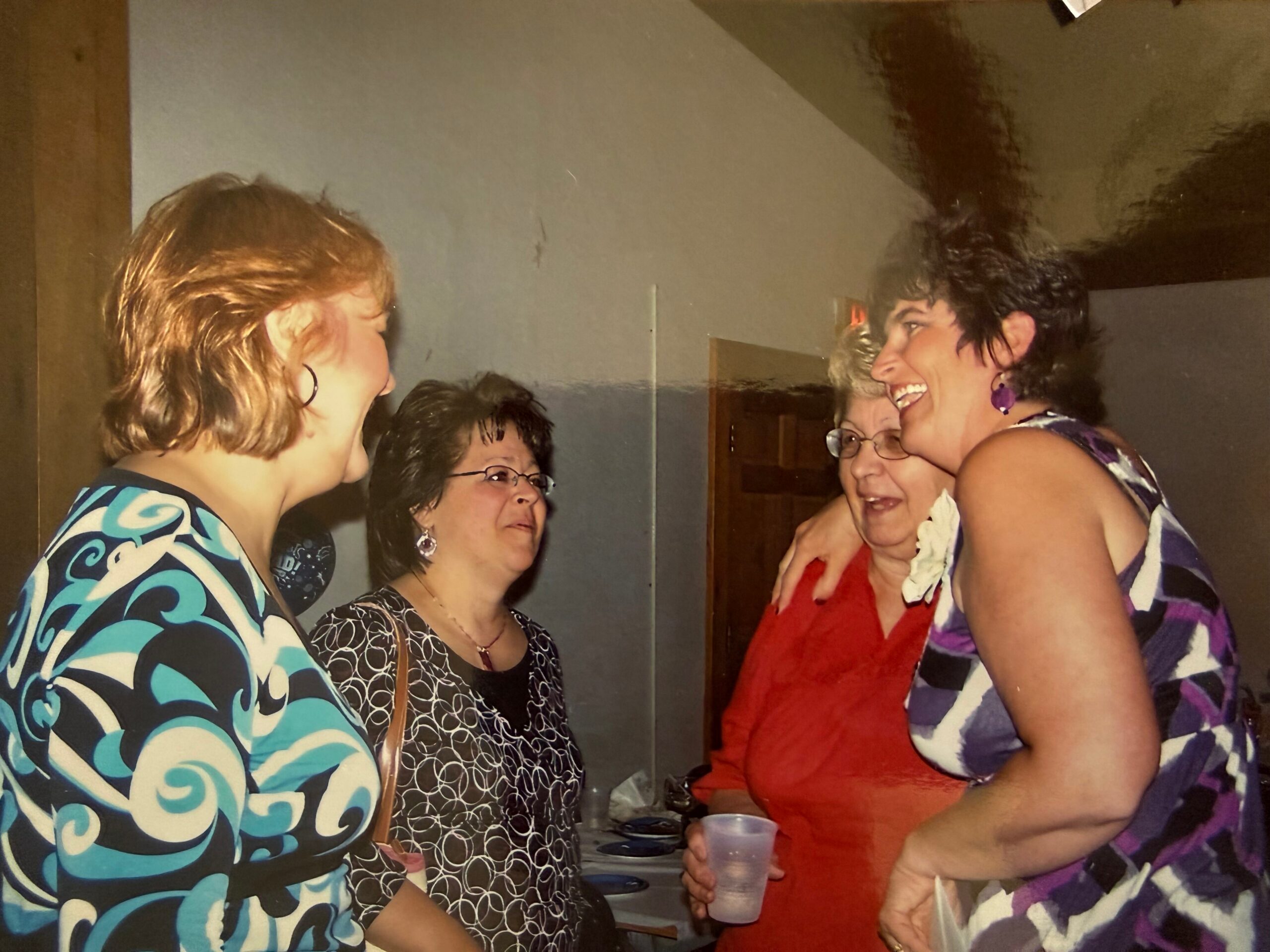Group of adults at a party indoors, smiling and chatting while holding plastic cups, wearing colorful dresses.