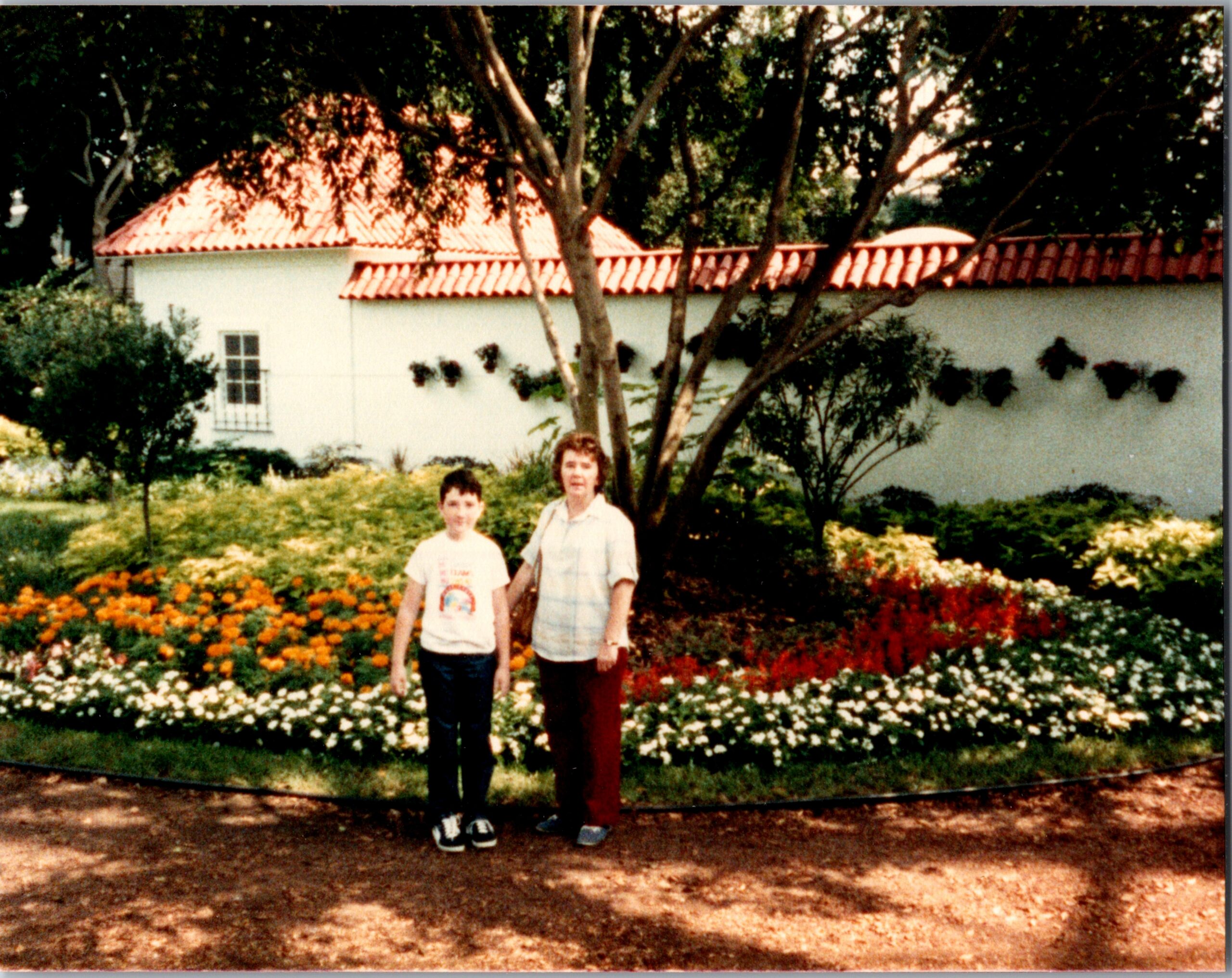 Boy and woman standing together in a flower-filled front yard, with a white wall, red-tiled roof, and trees behind them.