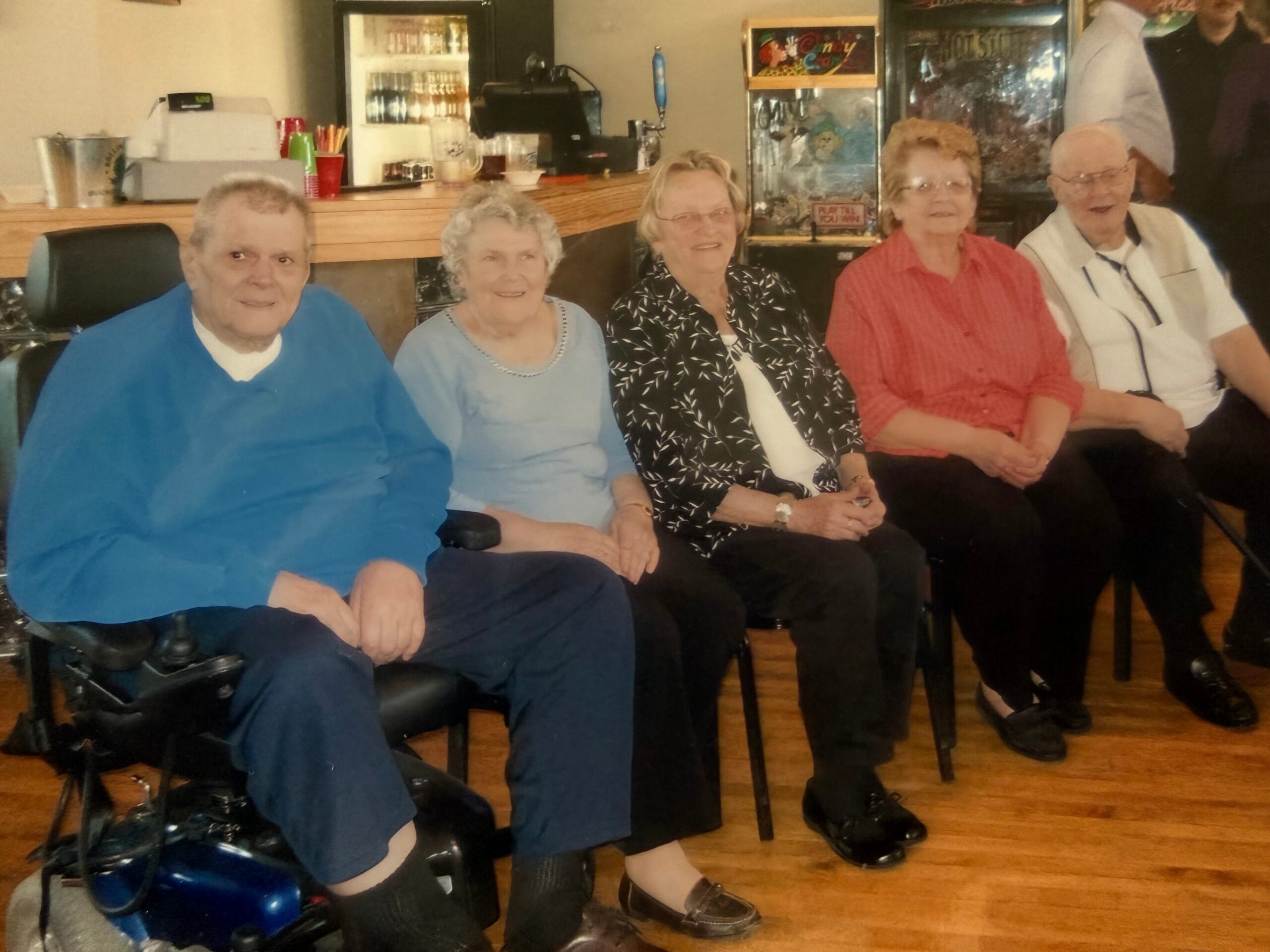 Five elderly friends seated together in a cafe, smiling at the camera, with a counter and arcade machines in the background.