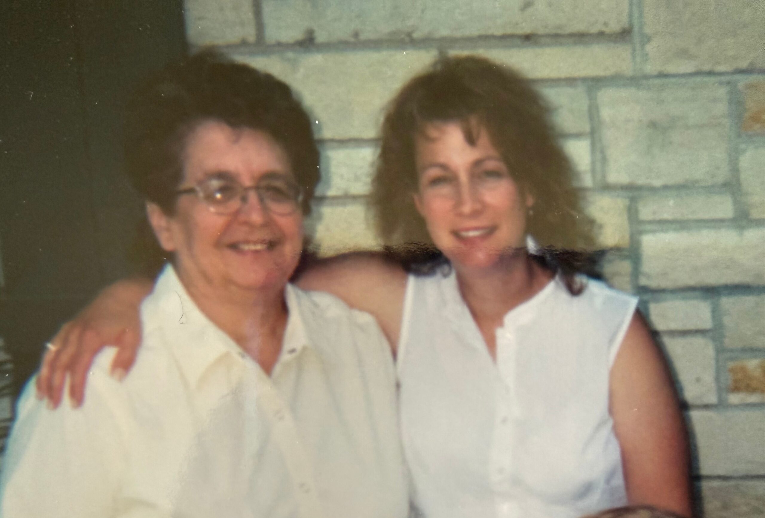 Two women smiling and posing closely together in light-colored sleeveless tops in front of a brick wall; appears to be an old, faded photo.
