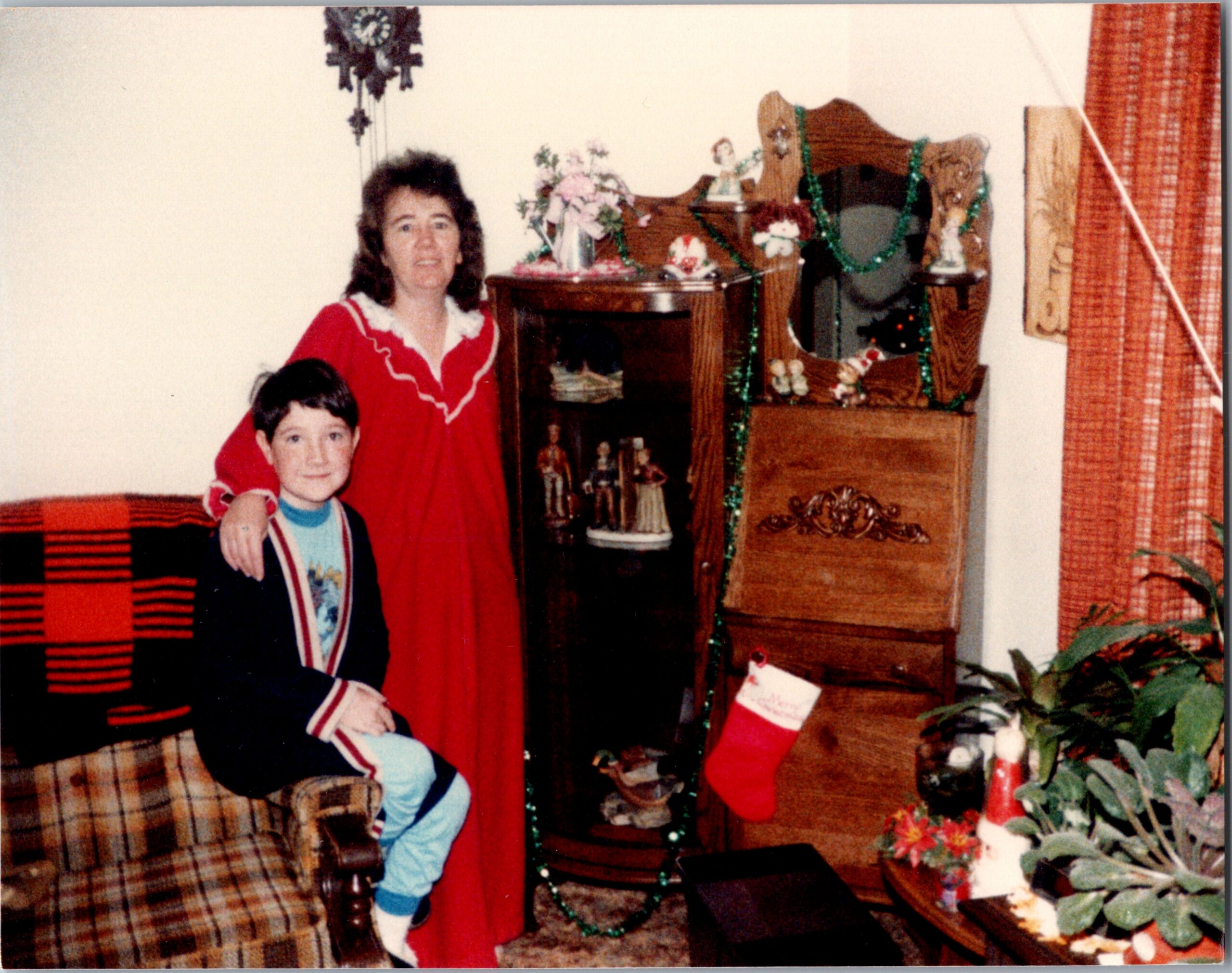 Woman in a red dress stands beside a boy in a holiday-decorated room with garlanded dresser and stockings dangling nearby.