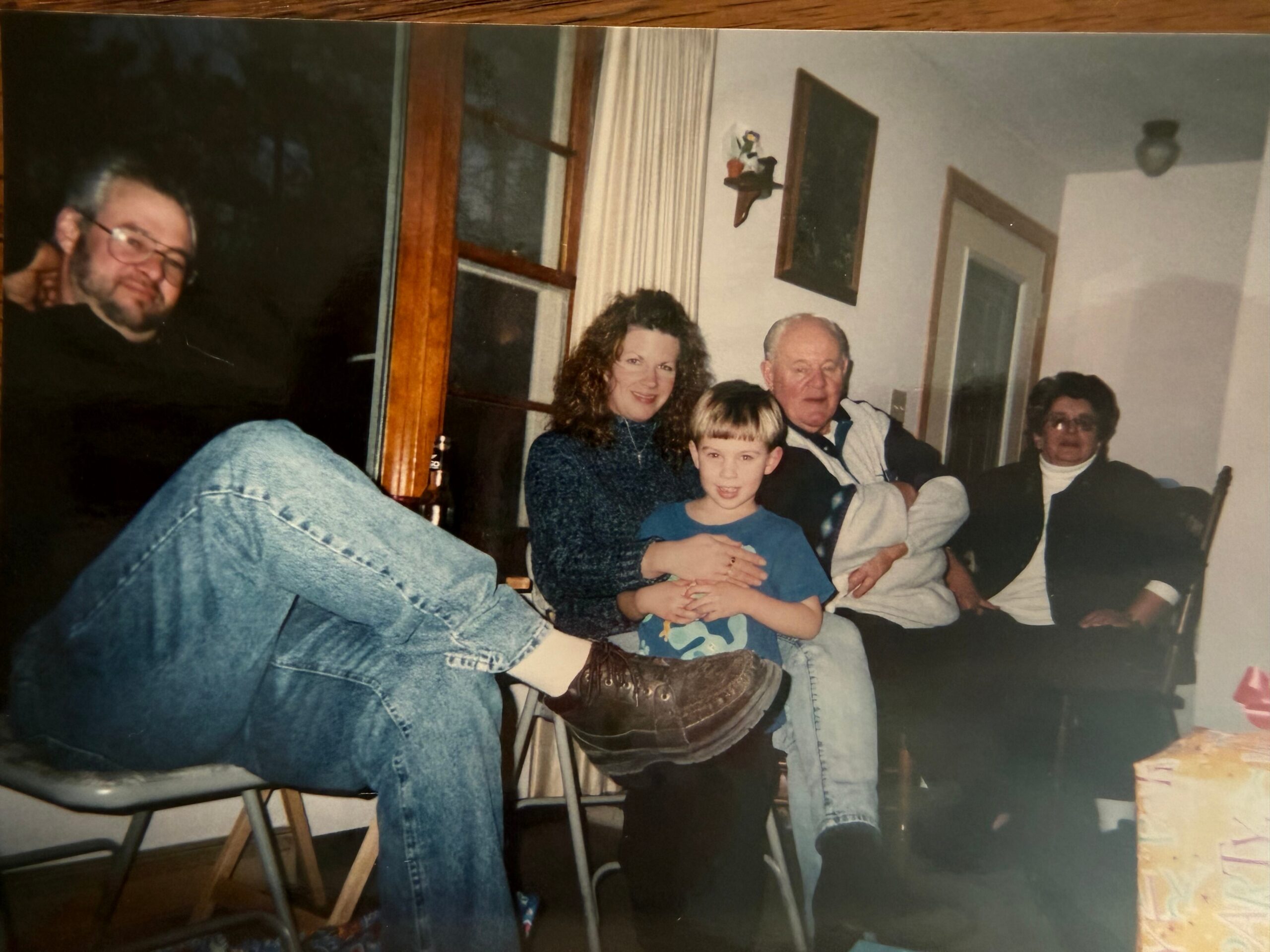 Family group posing for a casual photo in a living room, with a man in jeans on the left, a woman with curly hair, a young boy in blue, an older man, and another person seated on the right.