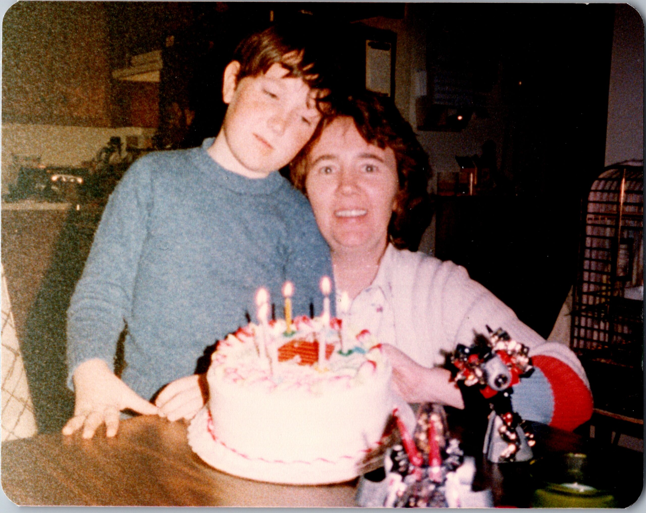 A young man and an older woman smile beside a multi‑candled birthday cake in a dim room.
