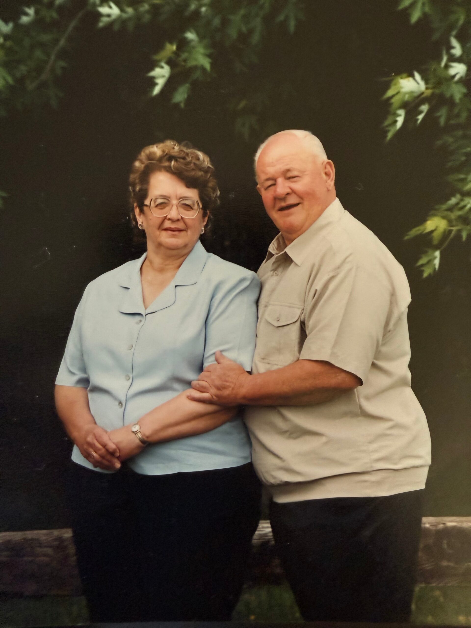 Older couple standing side by side outdoors, arms linked, smiling at the camera.