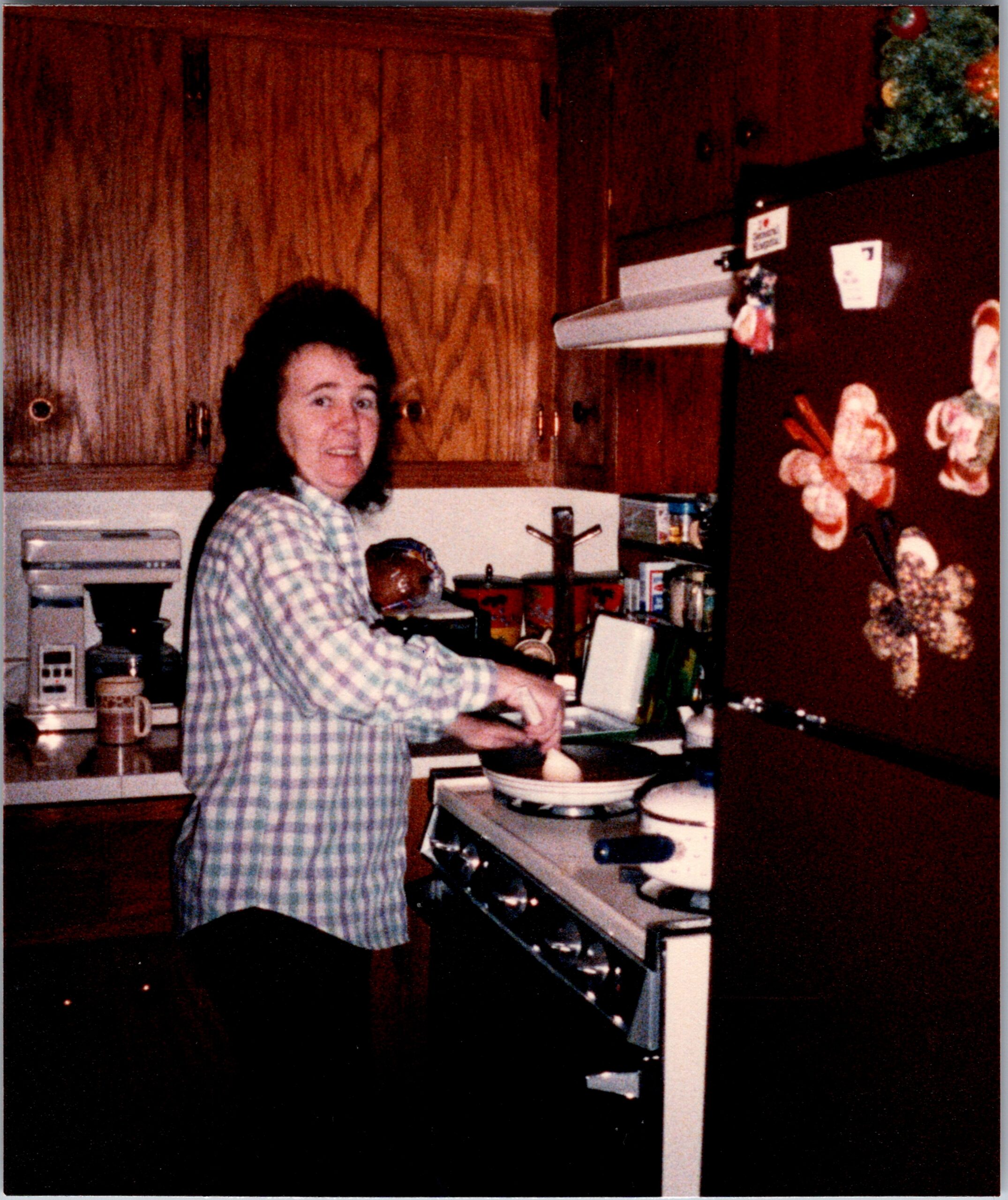 Woman in a blue plaid shirt cooking at a kitchen stove with wooden cabinets behind her.