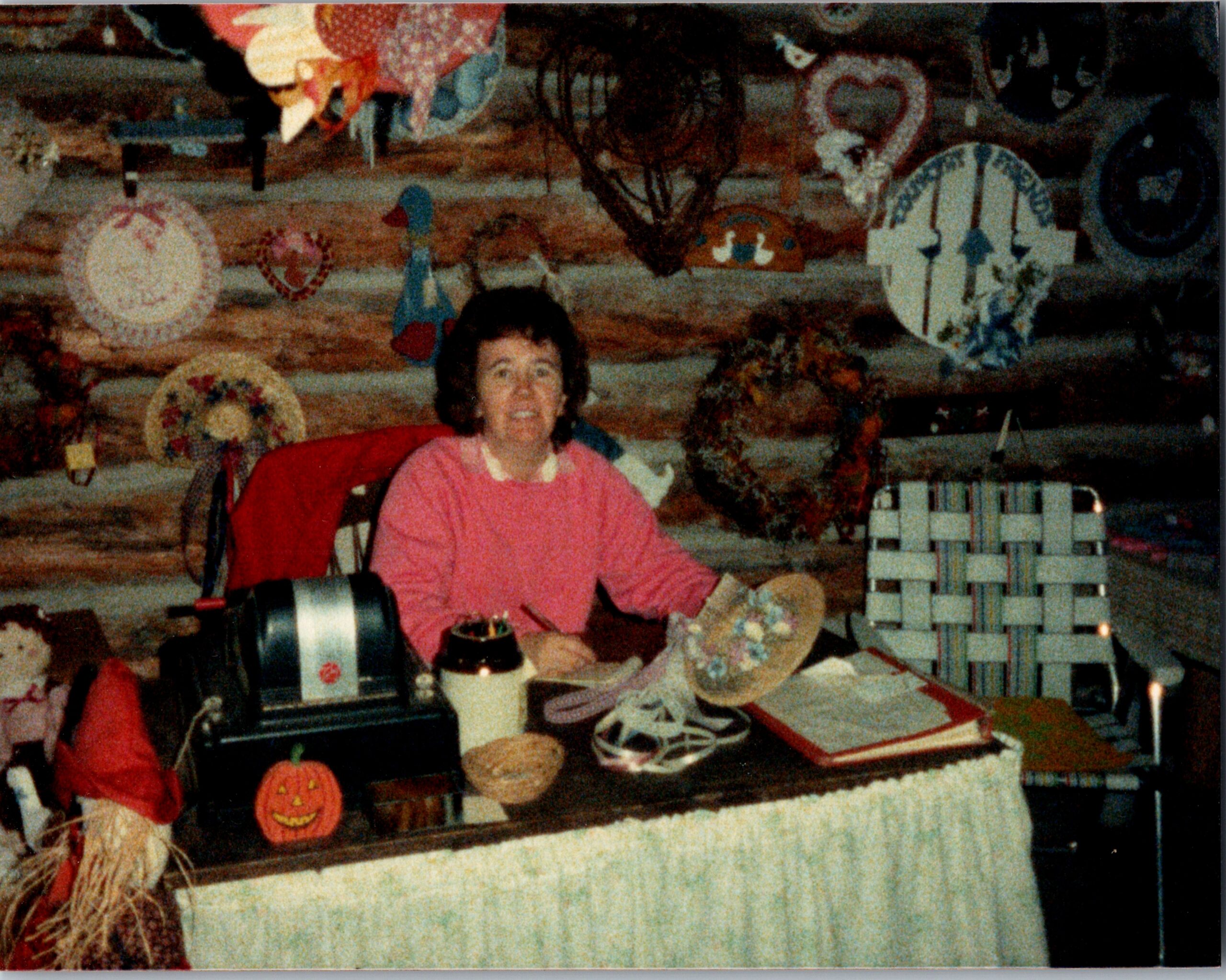 Smiling woman seated at a craft table with Halloween decorations, including pumpkins and handmade ornaments, in a cluttered workshop background.