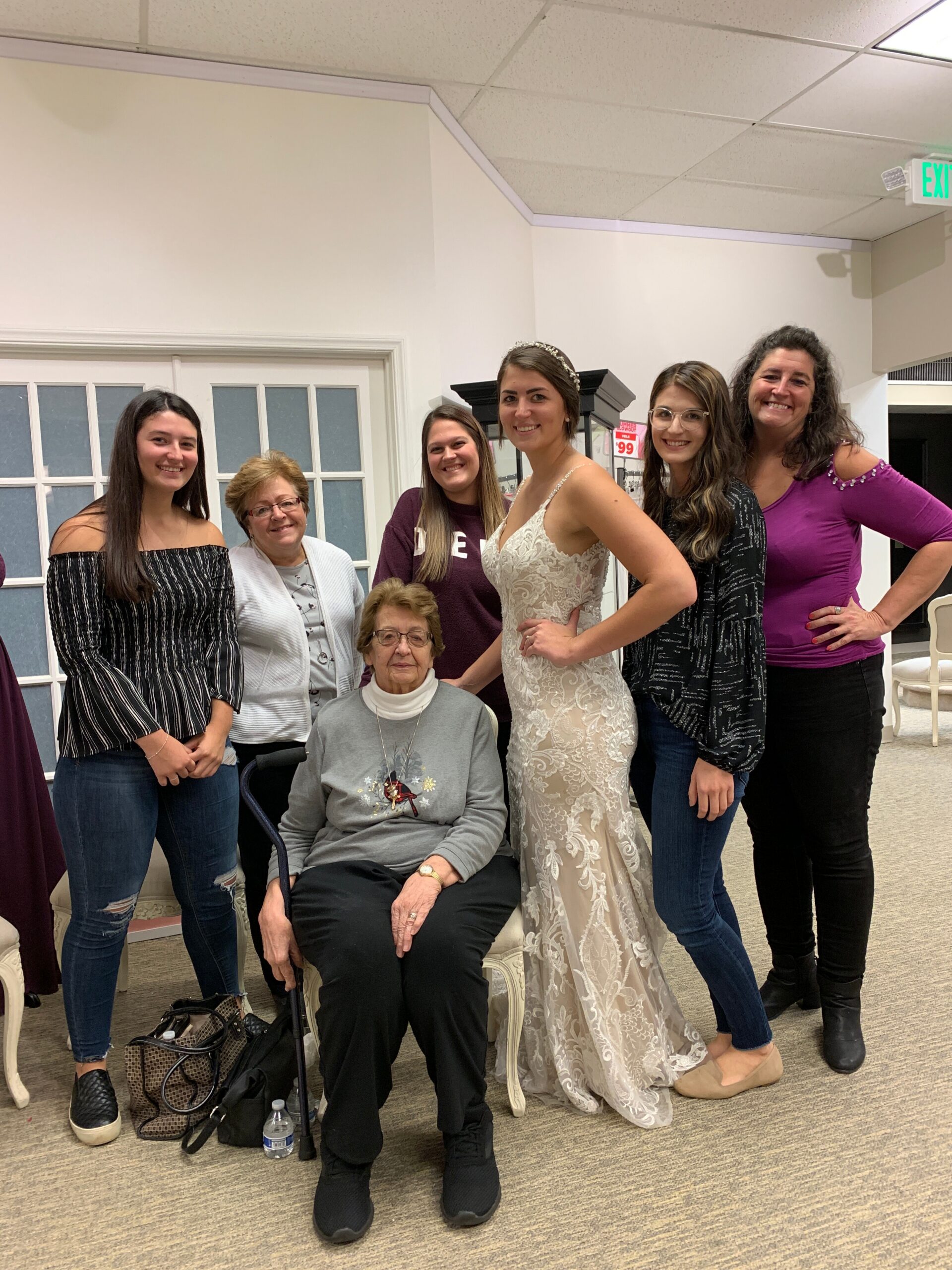 Bride in a lace wedding gown poses with friends and family indoors, one elderly woman seated in front next to her.