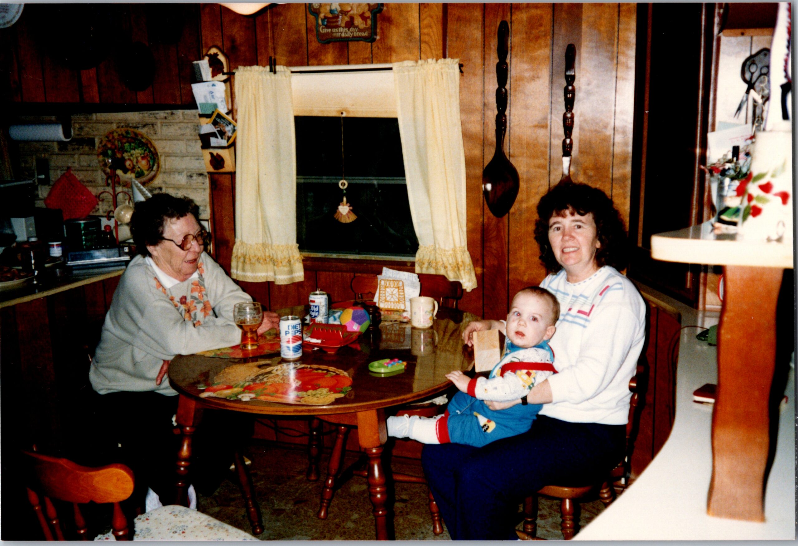 Grandma on the left at a wooden kitchen table with beverages, a mom holding a baby on the right, and a toddler at the table.”