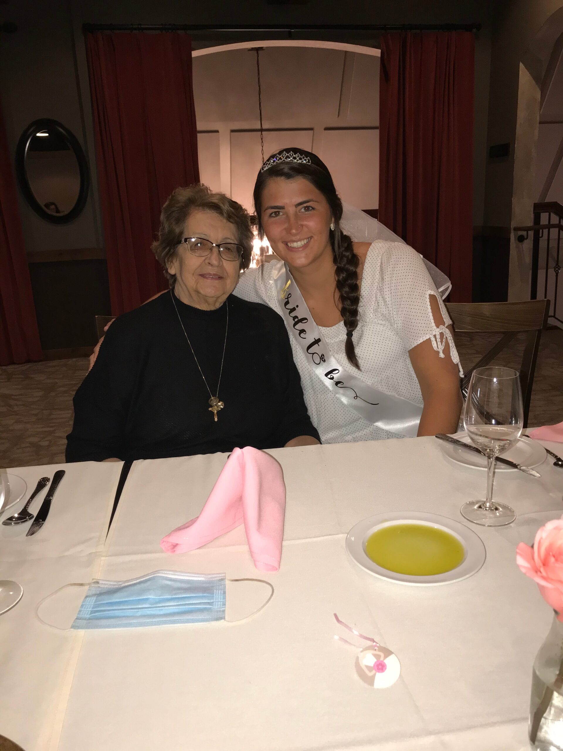 Two women smiling at a dining table; a younger woman wears a tiara and sash, seated beside an older woman in a black sweater in a restaurant setting.