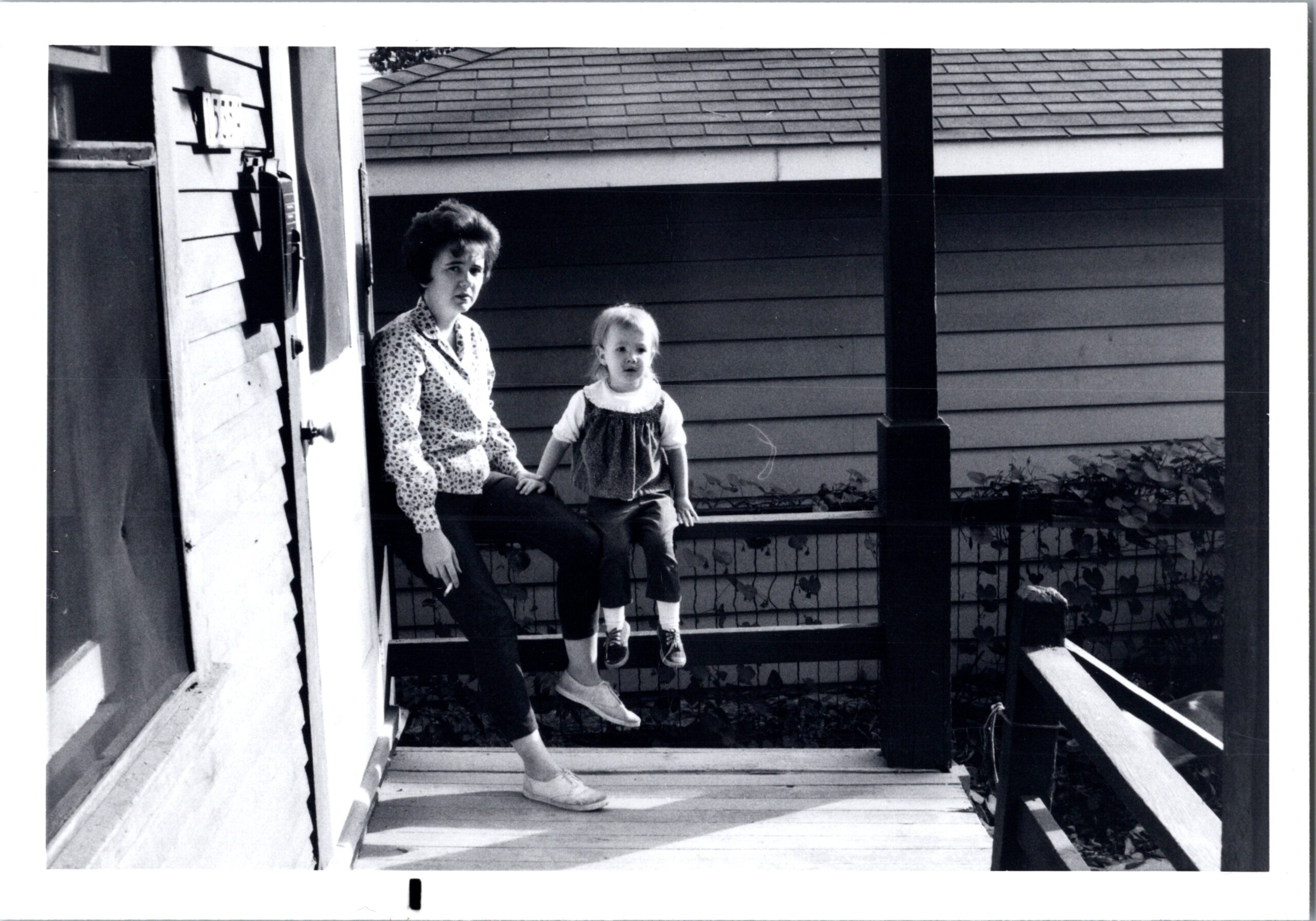 Woman and young child sitting on the front porch steps of a house, facing the camera; the woman wears a patterned blouse and dark pants while the child wears a dress overalls and sneakers.