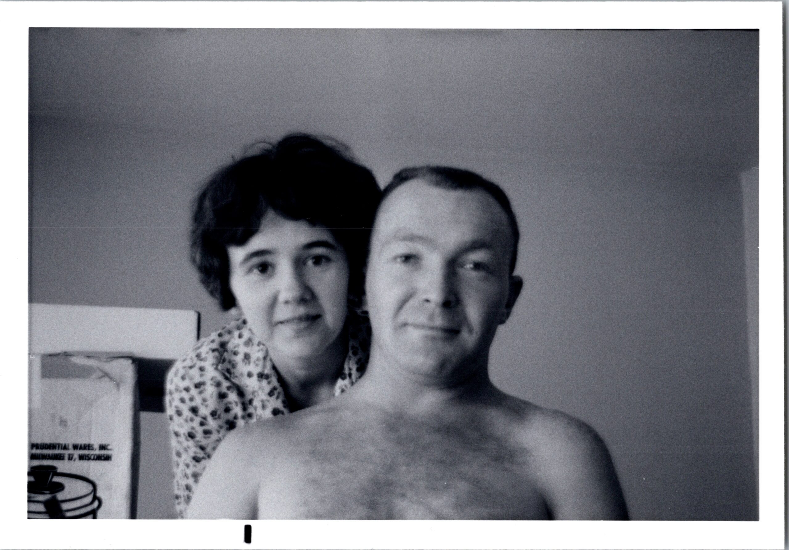Black-and-white photo of a smiling couple posing closely for a selfie, with the woman on the left wearing a patterned blouse and the man in front, shirtless.
