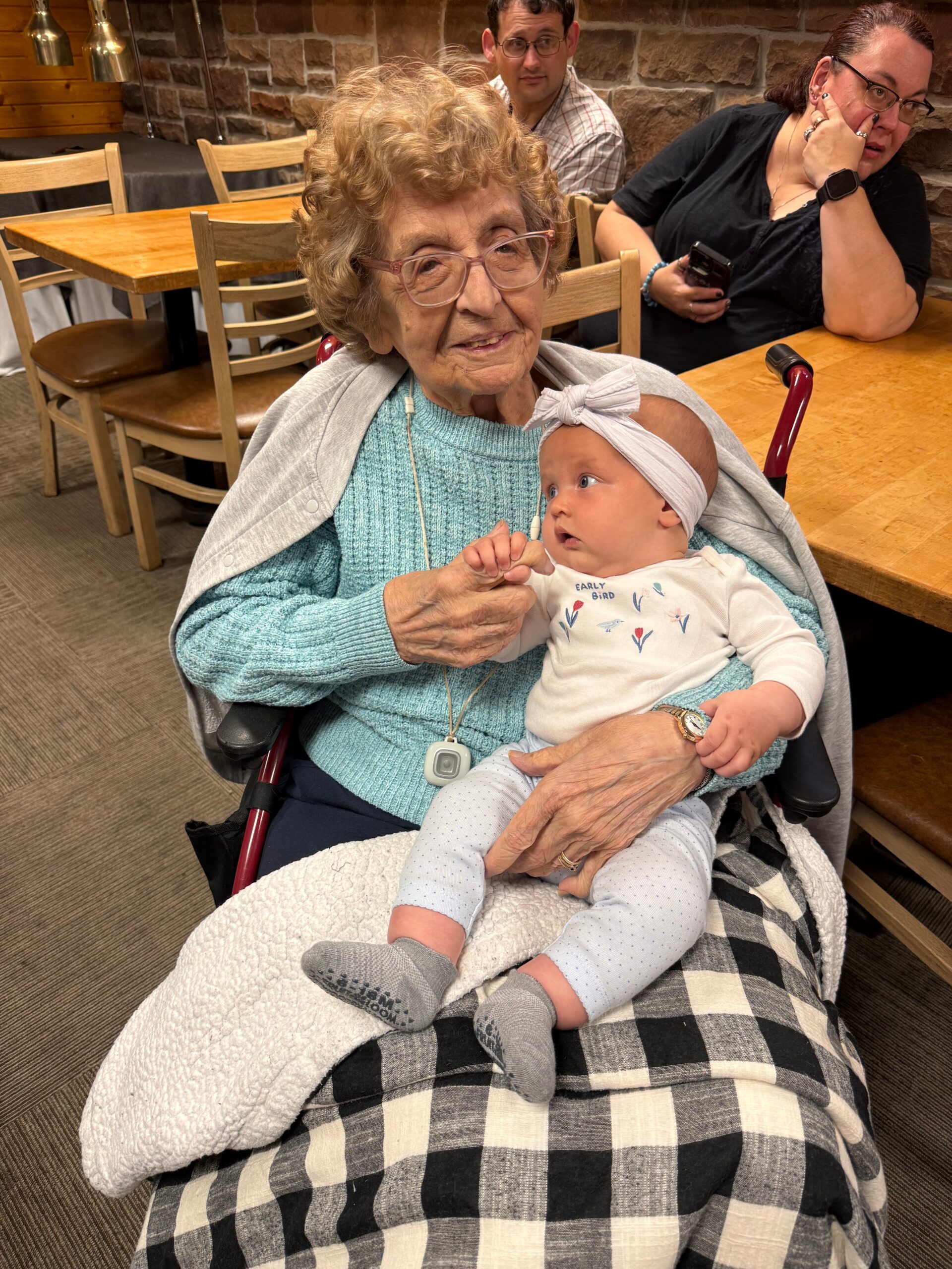 Elderly woman with glasses sits in a wheelchair, holding a baby wearing a headband, in a cozy restaurant setting.