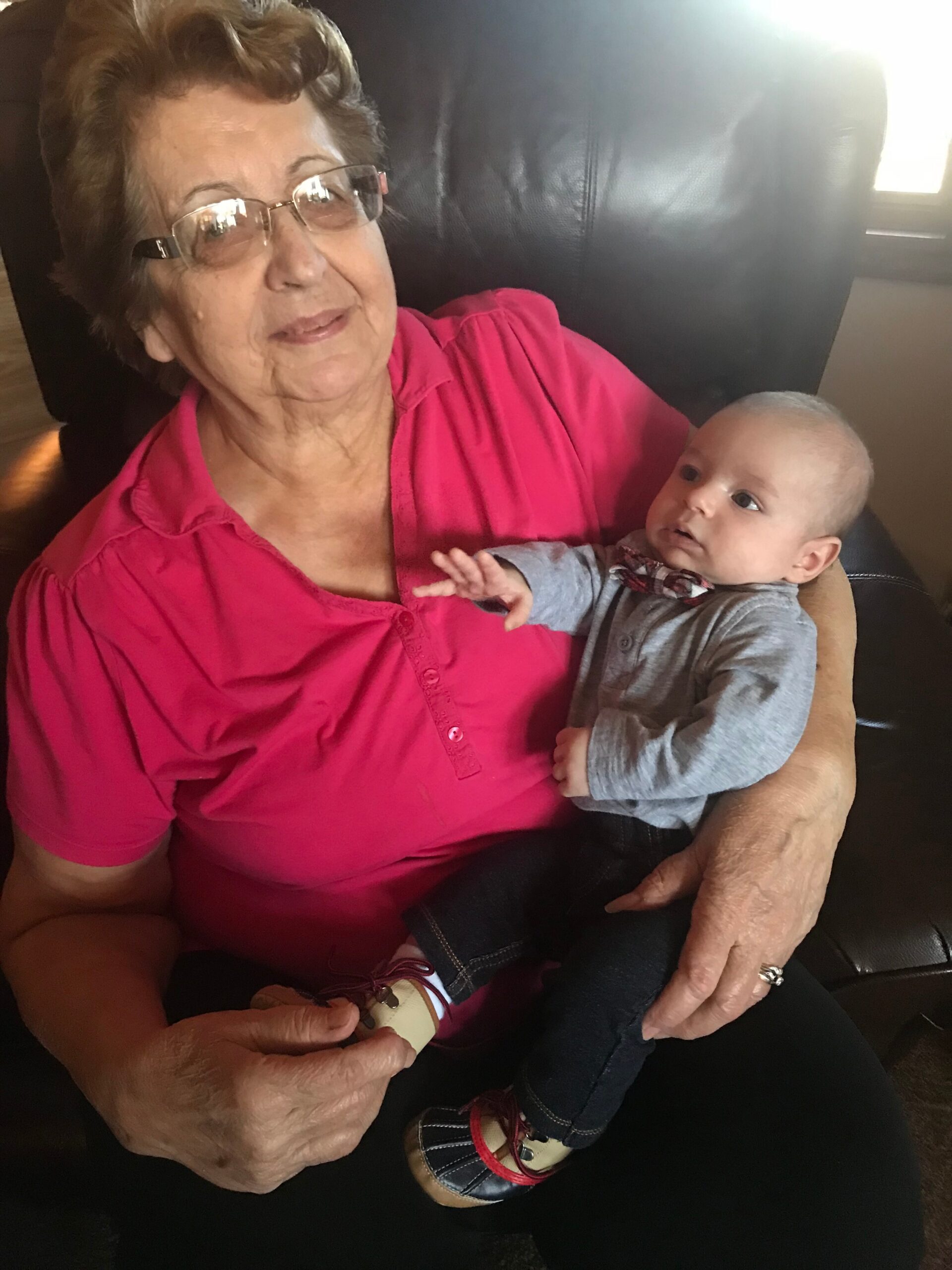 Grandmother in a pink blouse cradling a baby in gray clothing on a dark leather couch, both looking content.