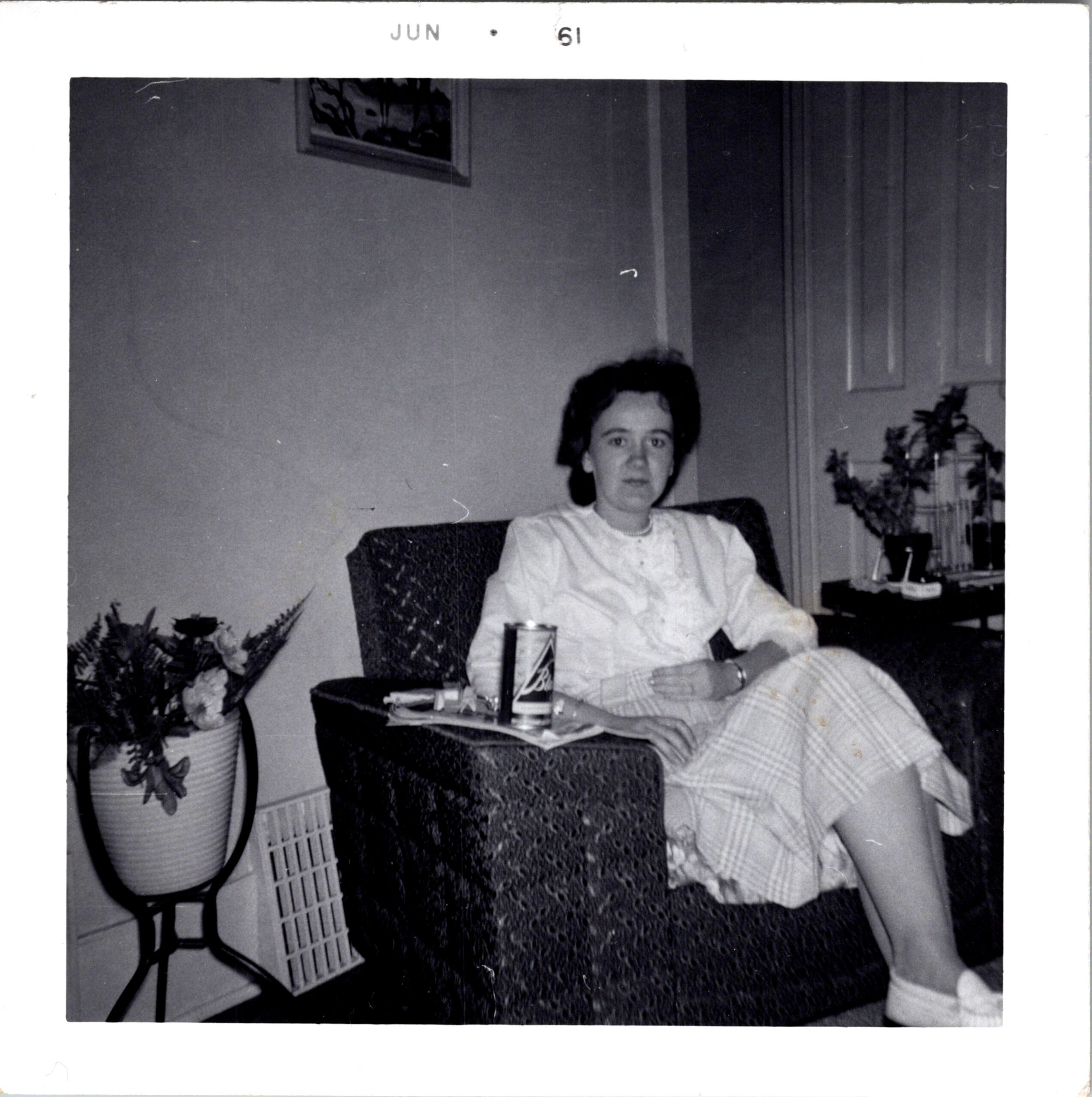 Woman seated in a living room chair, wearing a light blouse, with plants and a cabinet in the background.