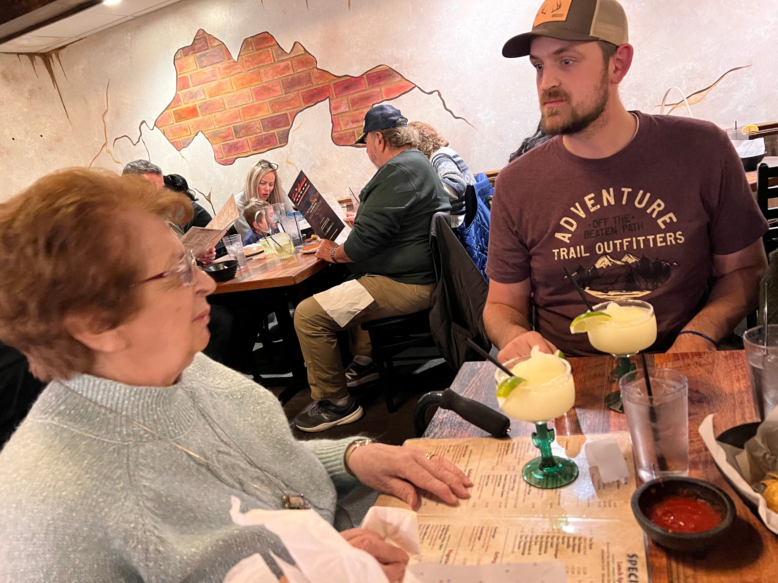 People seated at a long wooden table in a restaurant, with drinks and menus visible; mural brick wall in the background.