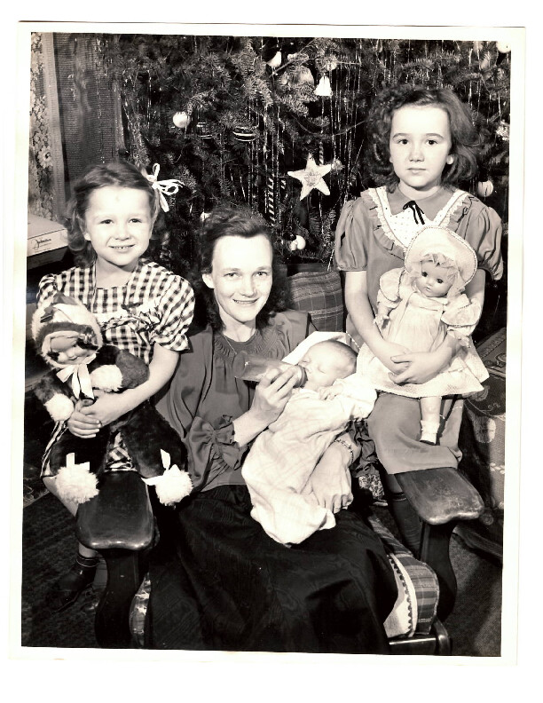 Woman seated center holding a newborn baby, flanked by two girls with toys, in front of a Christmas tree.