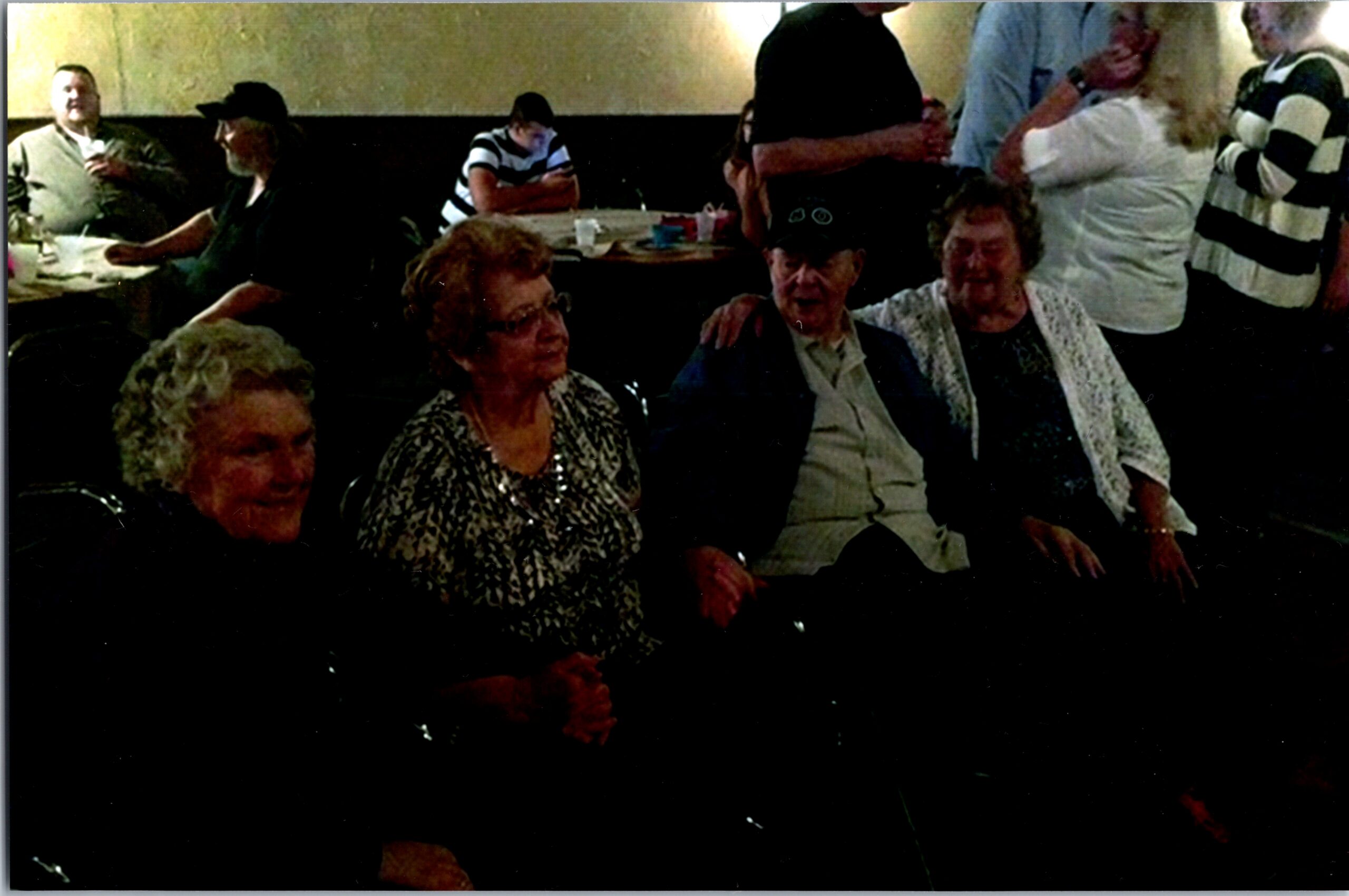 Group of seniors seated around tables in a dim restaurant, chatting and smiling at a social gathering.