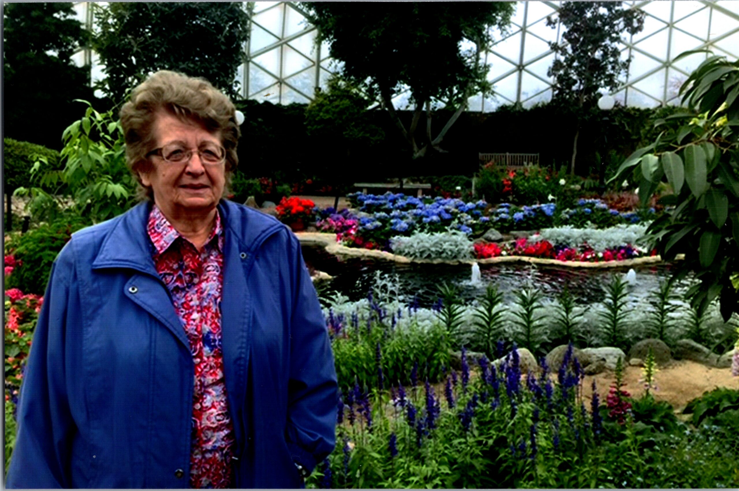 Older woman in a blue jacket standing in a colorful garden with a pond and flowers behind her.