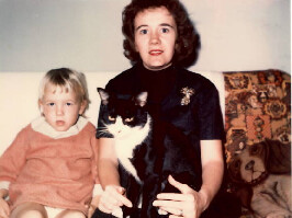 A woman sits with a toddler on her left and a black-and-white cat on her lap in a living room portrait.