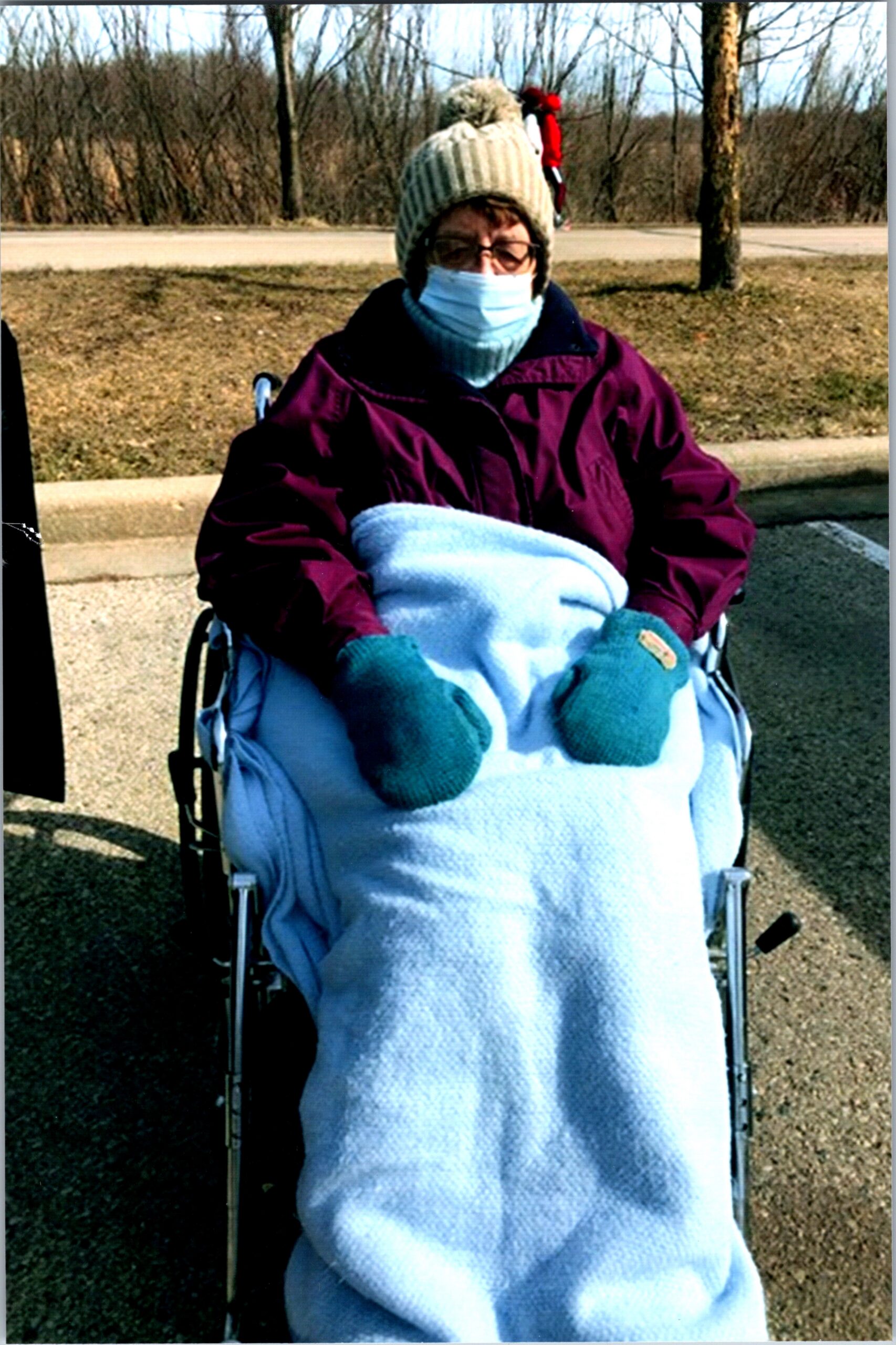 Person in a wheelchair outdoors on a cold day, wrapped in a light blue blanket with a burgundy jacket, teal mittens, mask, and knit hat.