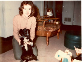 Woman kneeling on a carpet, holding a black dog with a ribbon in a living room.