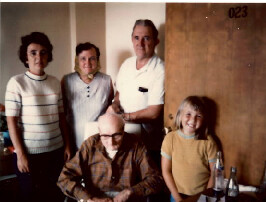 Five adults in a home setting pose for a photo: one man sits at a table signing papers while four others stand behind him, smiling.