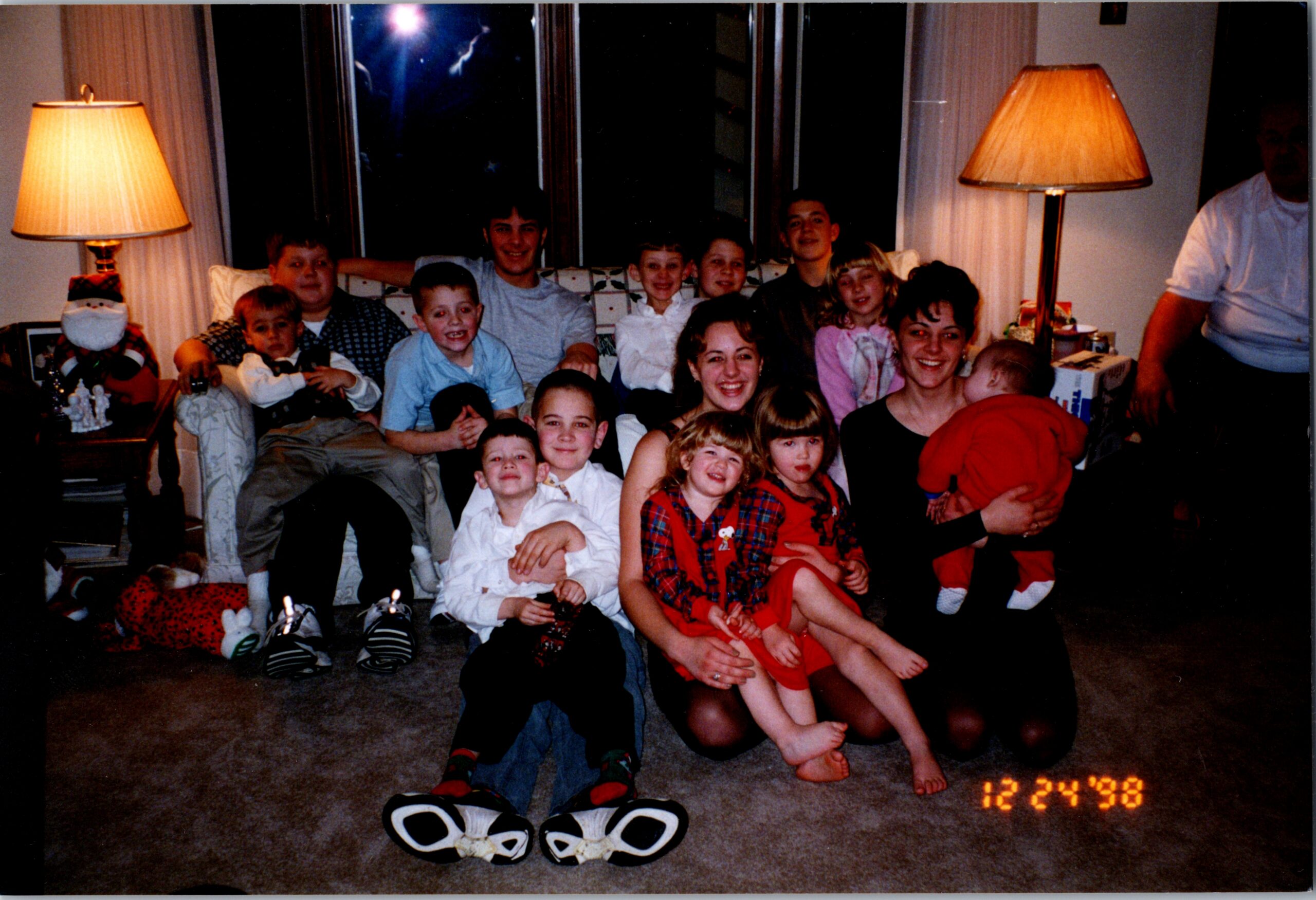 Family Christmas photo: many children and adults smiling together in a living room, sitting on a couch and floor with holiday decor and a Santa figure; date stamp 12/24/98.