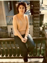Woman sits on a wooden railing on a sunny porch, wearing a peach sweater and dark jeans.