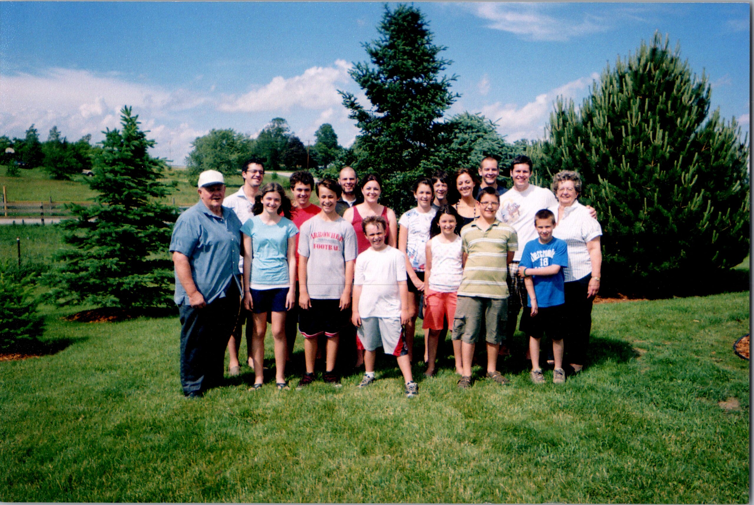 Group of people posing outdoors on a sunny day, standing on a grassy lawn with trees and a blue sky in the background.