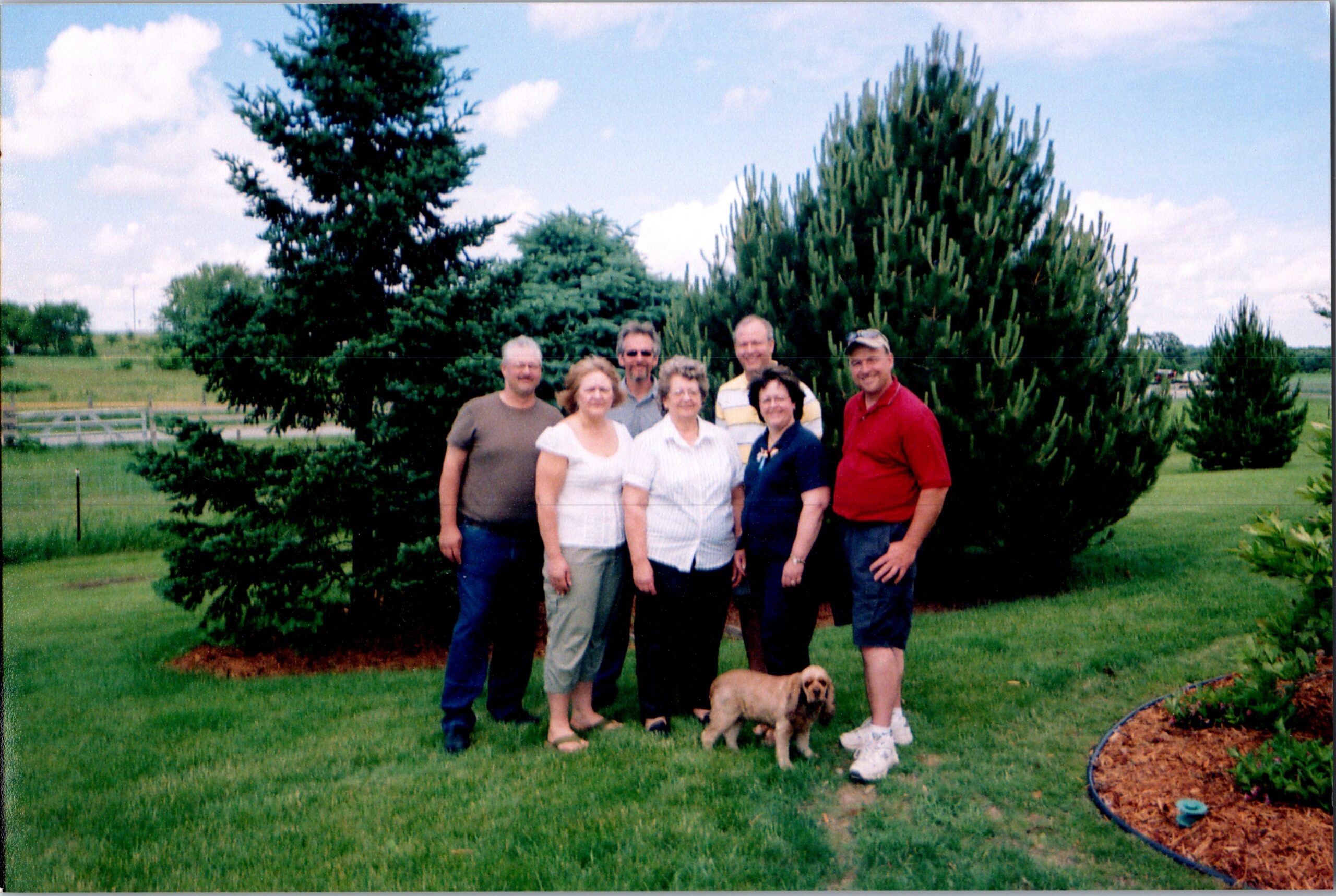 Group of seven adults posing together outdoors beside tall evergreen trees on a sunny day, with a small dog in front.