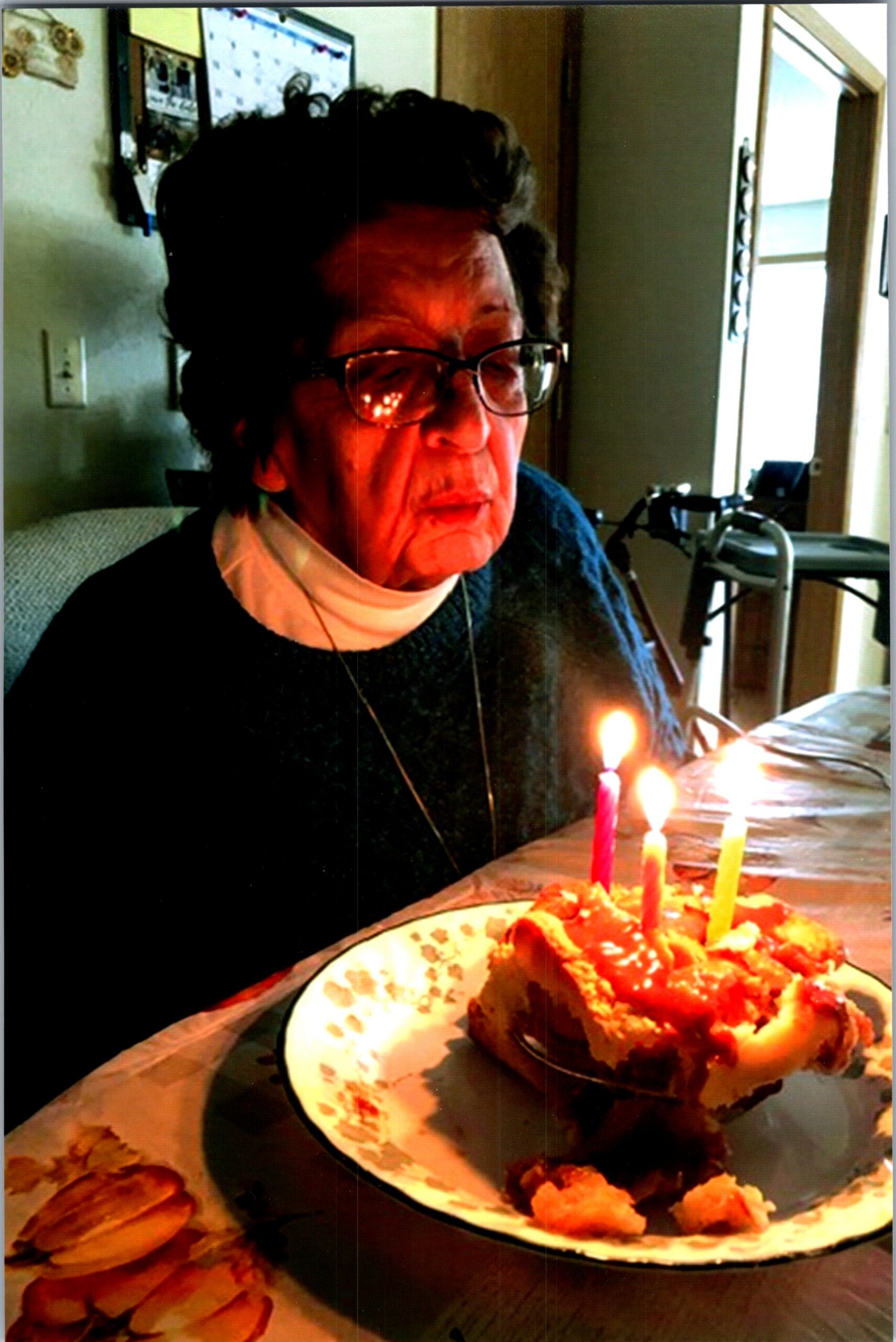 Older woman with short dark hair and glasses sits at a table, looking toward a frosted birthday cake with lit candles.