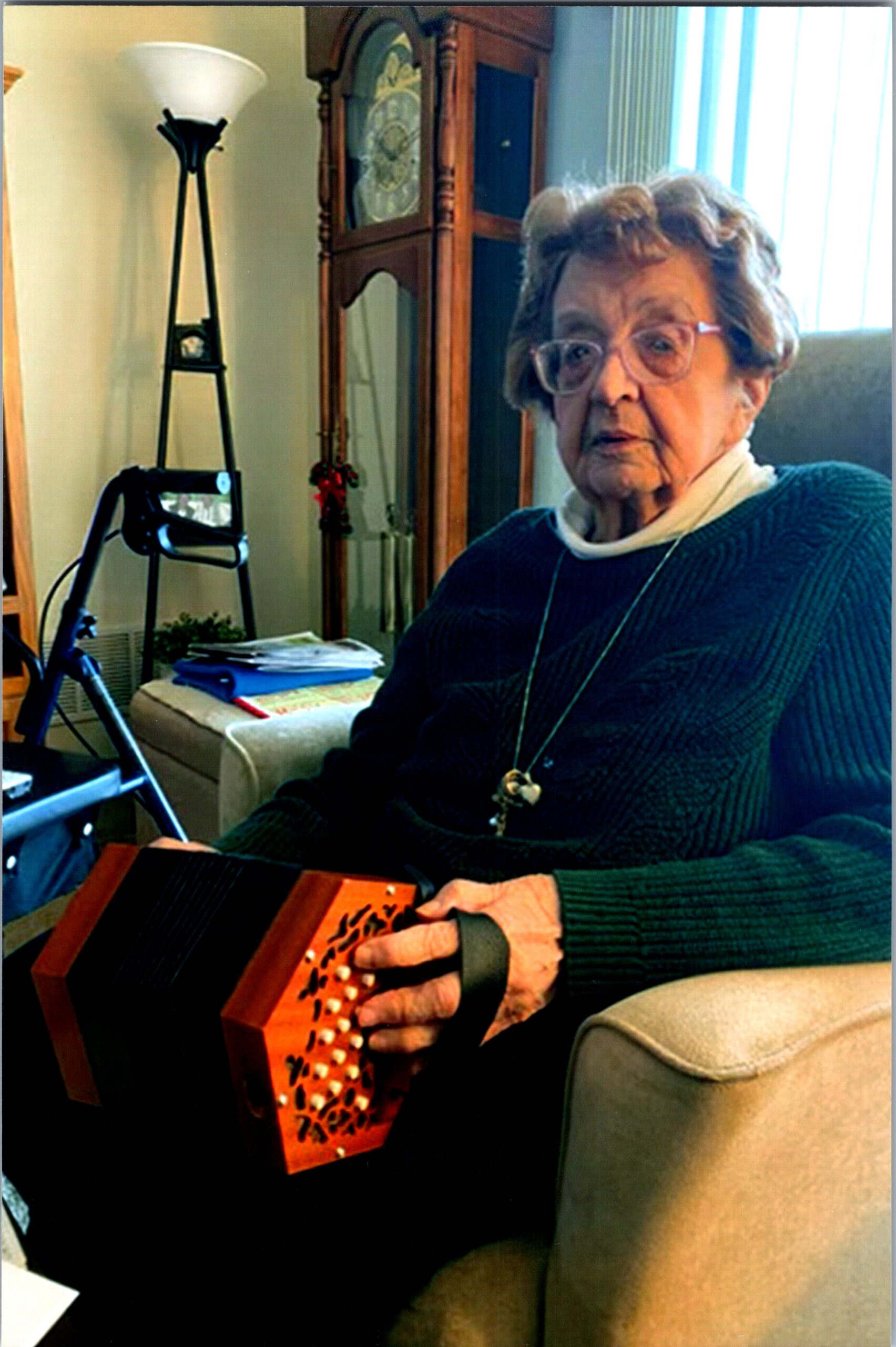 Elderly woman in a green sweater sits on a couch, playing a wooden hammered dulcimer in a living room.