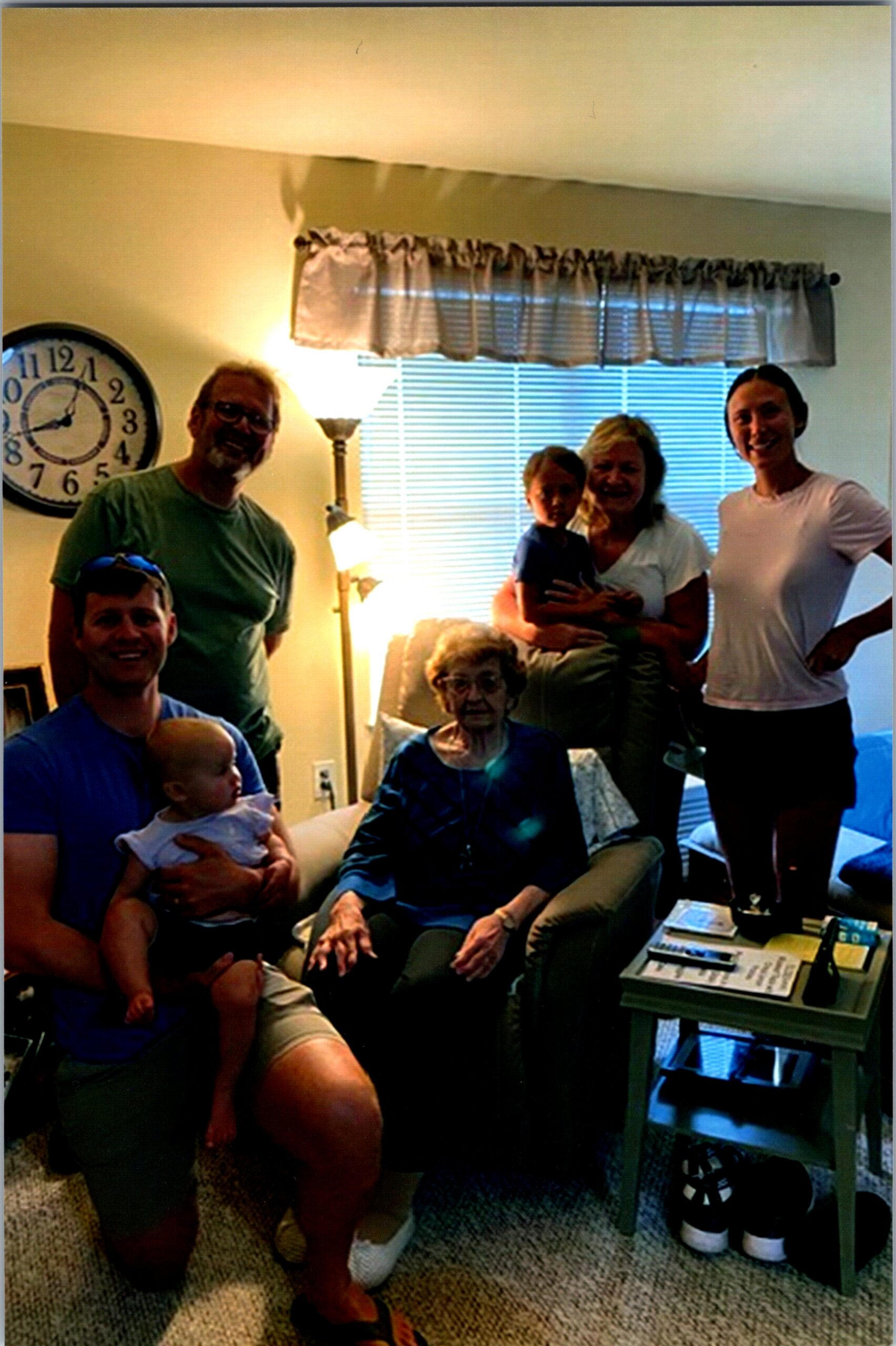 Family group photo in a living room: elderly woman seated, several adults standing around, and a baby held by a man; clock on wall behind them and window blinds in view.