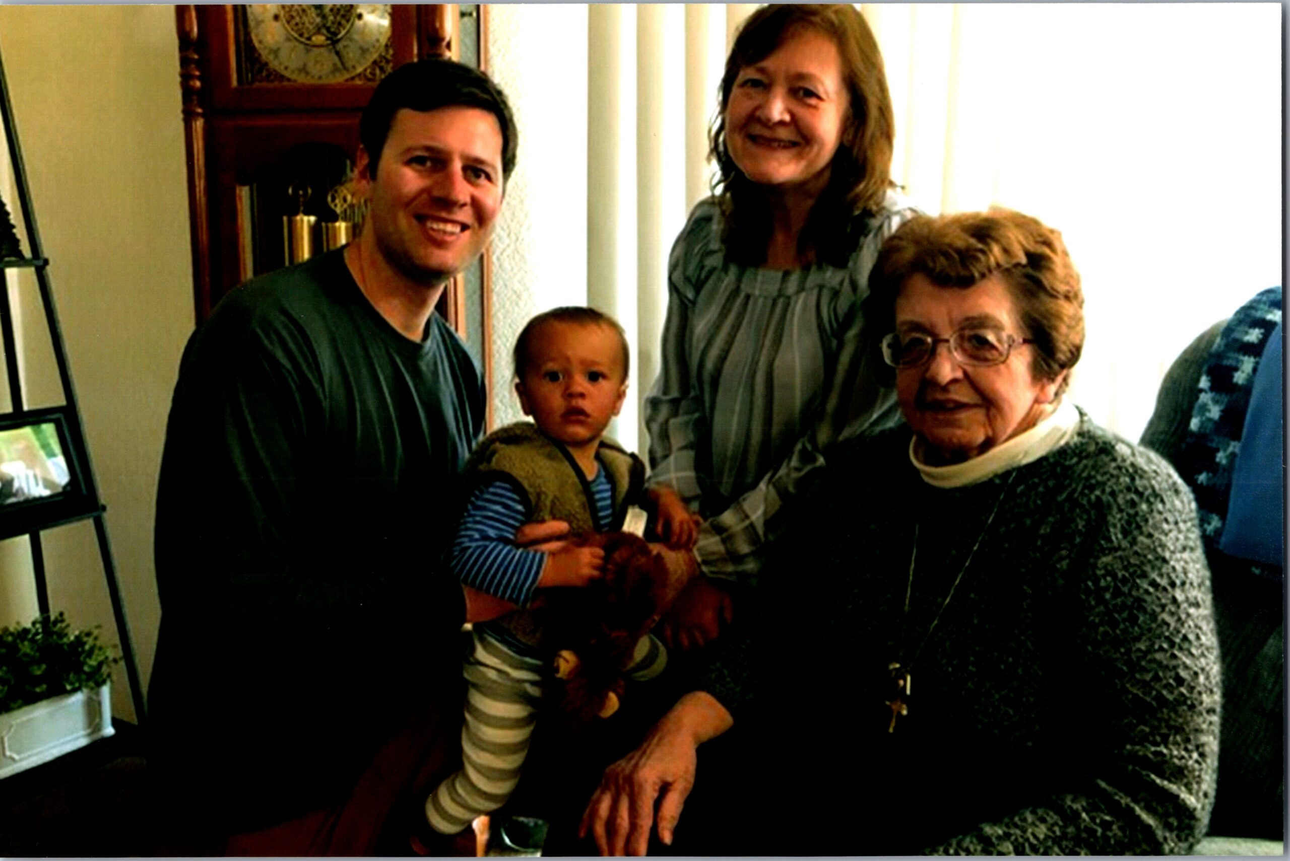 A family of four posing indoors: man holding a baby, a woman standing behind, and an older woman seated, with a tall clock in the background.