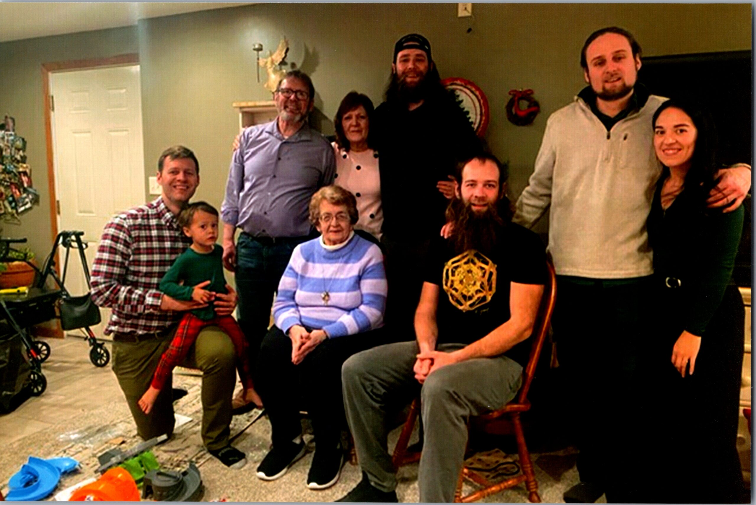 Group photo of family and friends in a living room, some seated and others standing, with a stroller and scattered toys on the floor and holiday decorations in the background.