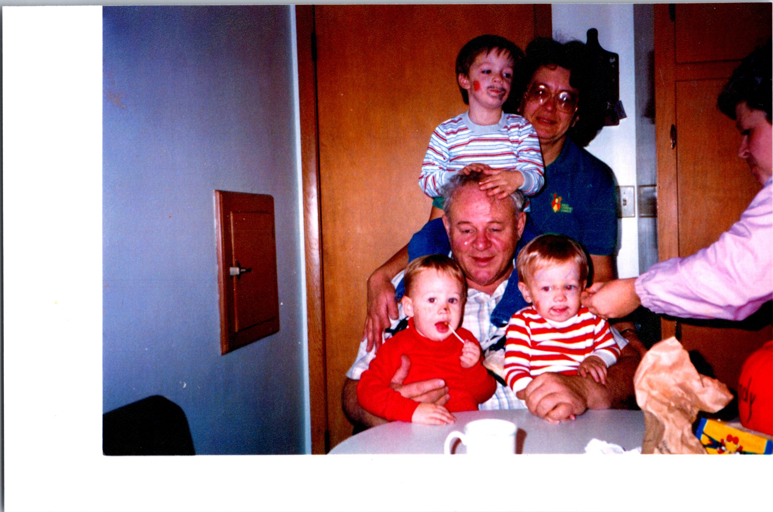 Man holds a young boy on his shoulders as several children sit at a table in a kitchen during a family gathering.