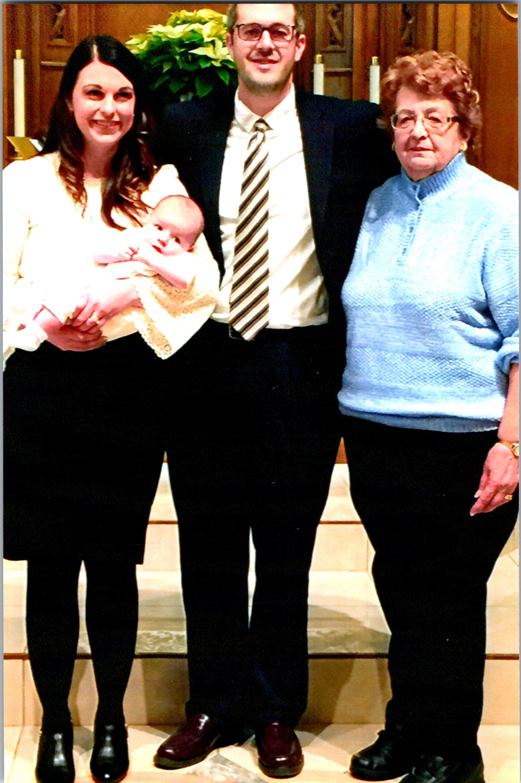 Family group posing indoors: woman holding a baby, man in a suit and tie, and an older woman in a blue sweater, all smiling.