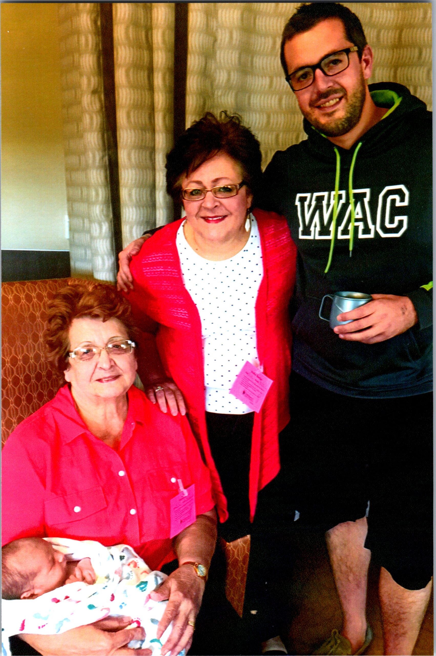 Group photo of three people indoors: an elderly woman in red, a middle-aged woman in a polka-dot white top, and a man in a black WAC hoodie with glasses, holding a mug; a baby is in a blanket in the foreground.