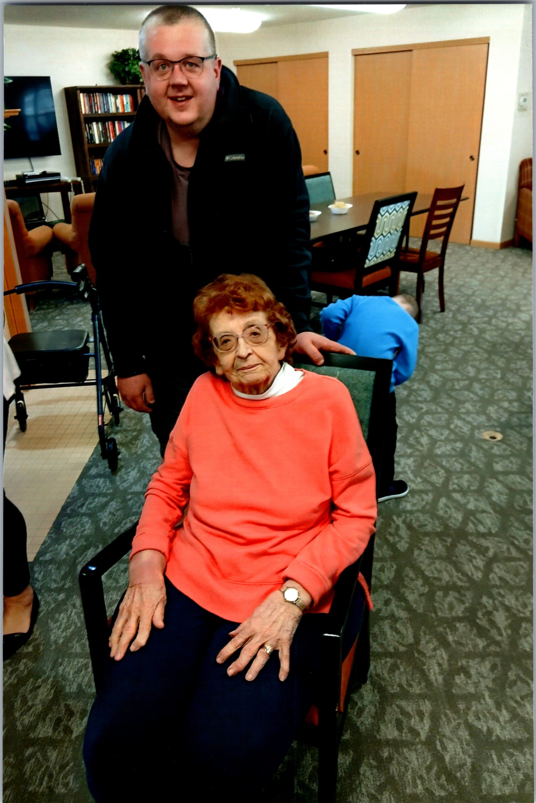 Older woman in a coral sweater sits in a wheelchair in a communal room, with a smiling man standing behind her.