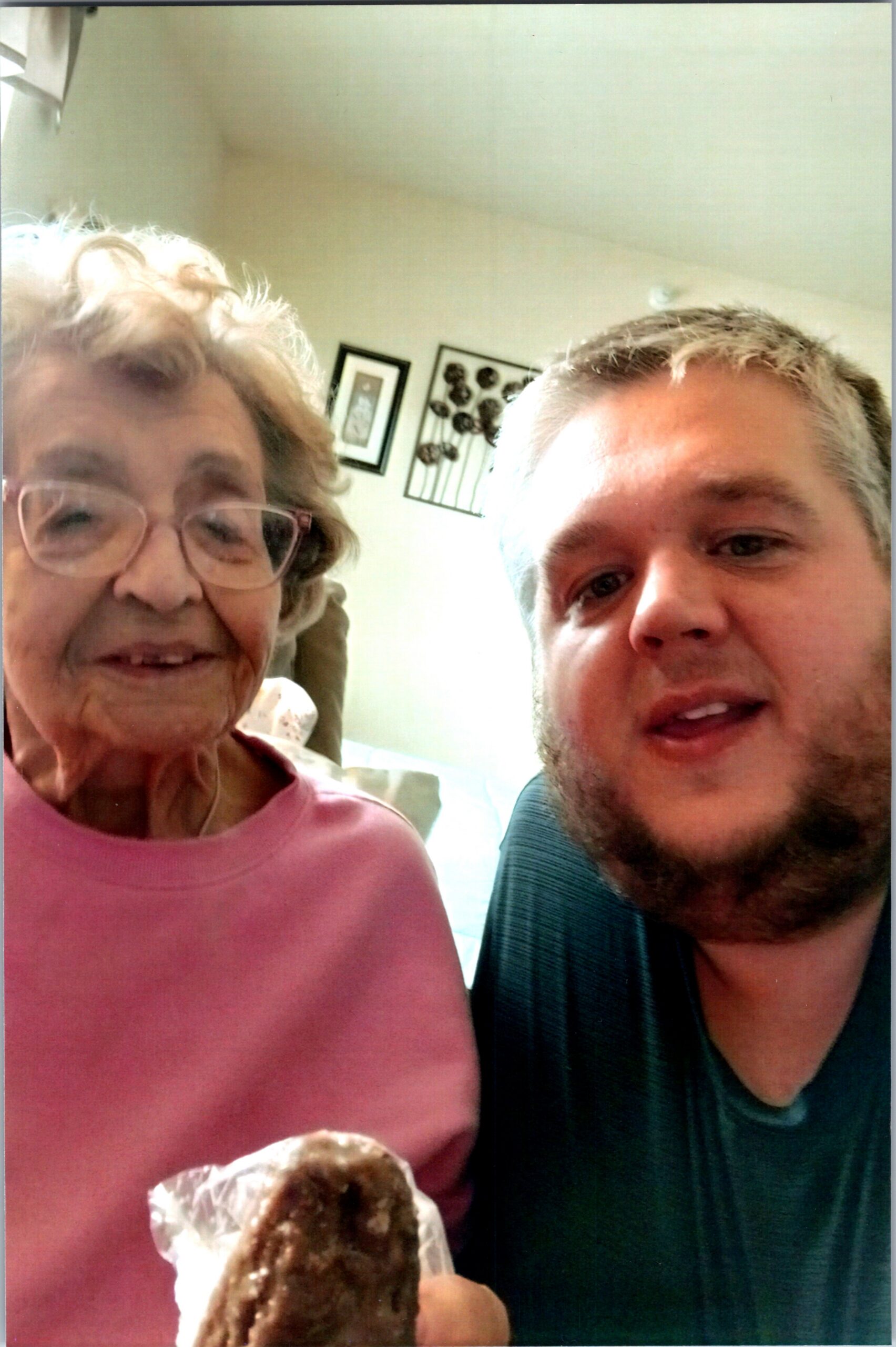 Close-up selfie of an elderly woman and a younger man smiling, each holding a chocolate donut wrapped in foil.