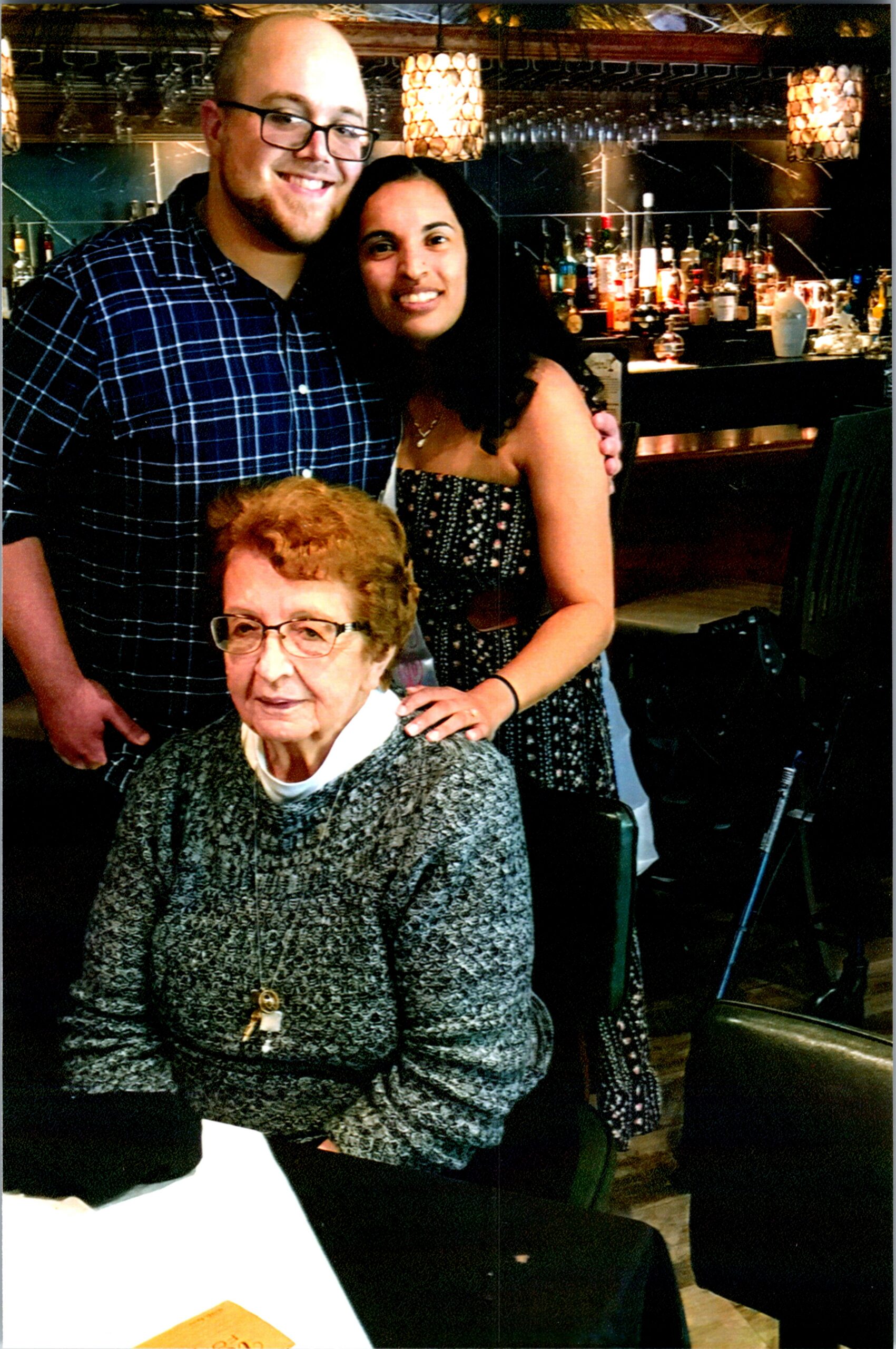 Older woman seated in front, with a smiling man in a blue plaid shirt and a woman in a strapless dress behind her, in a bar.