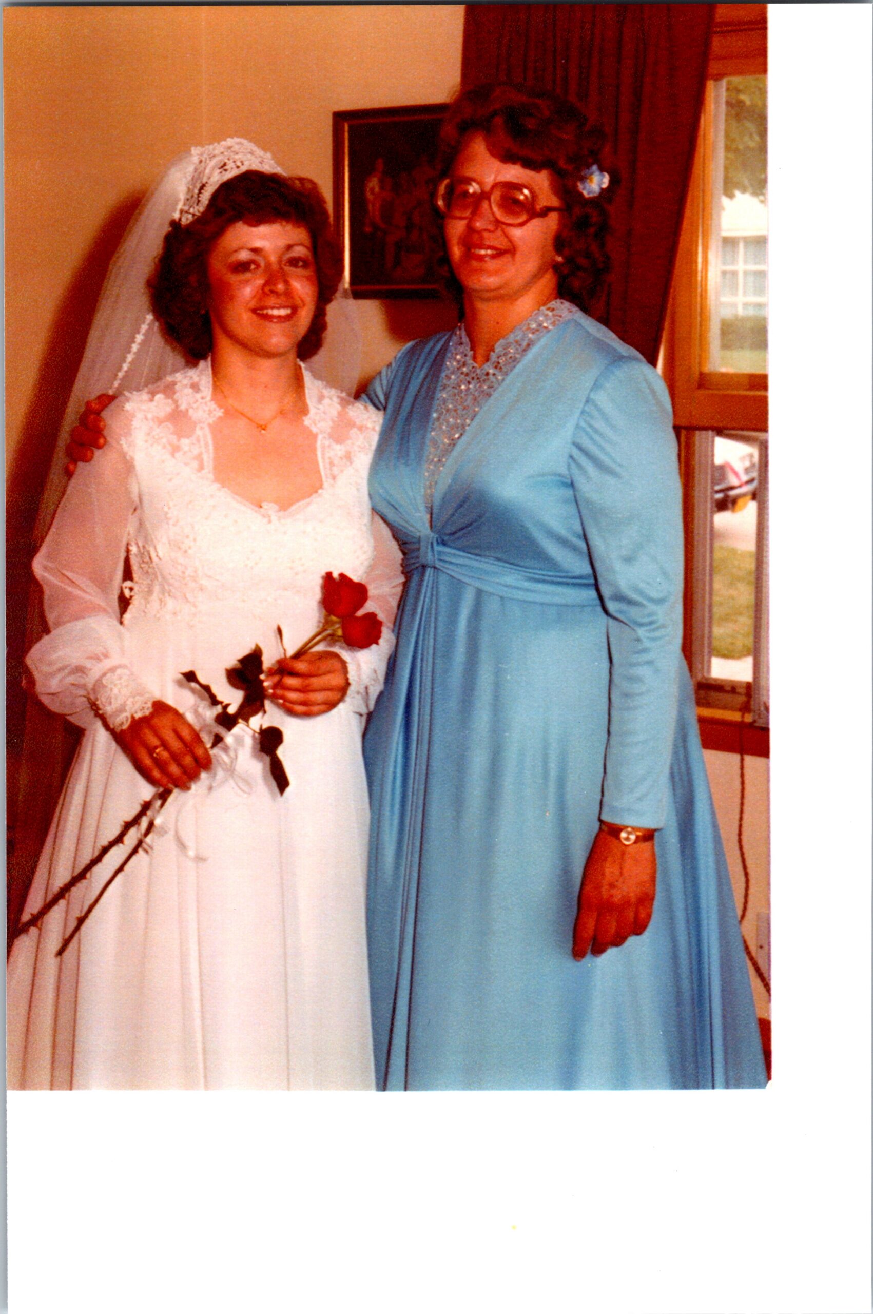 Bride in a white lace wedding dress and veil holding roses, posing with a guest in a blue dress indoors near a window.