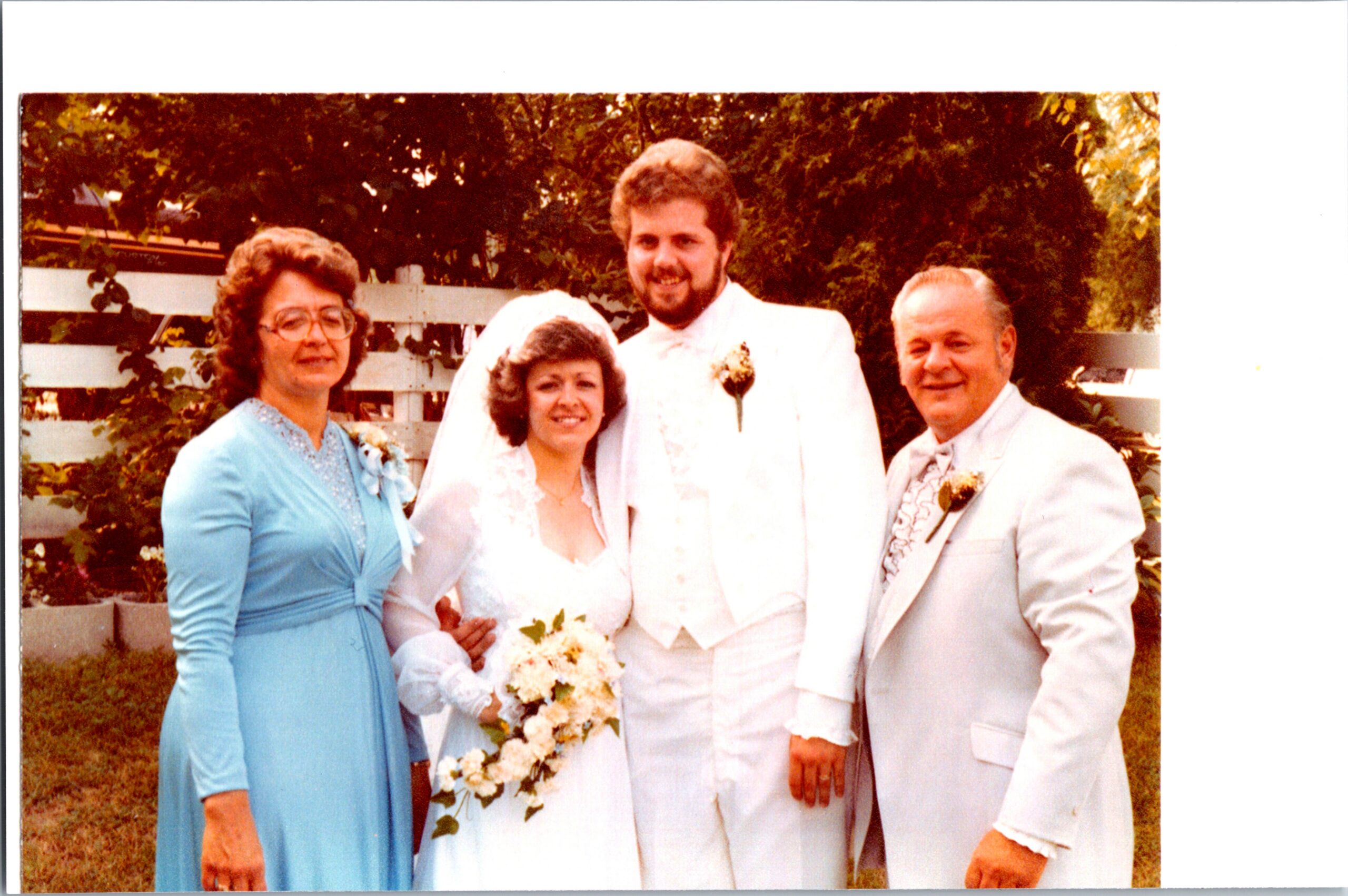 Bride in a white wedding gown holding a bouquet, standing with groom in a white suit and two guests outdoors by a white fence.